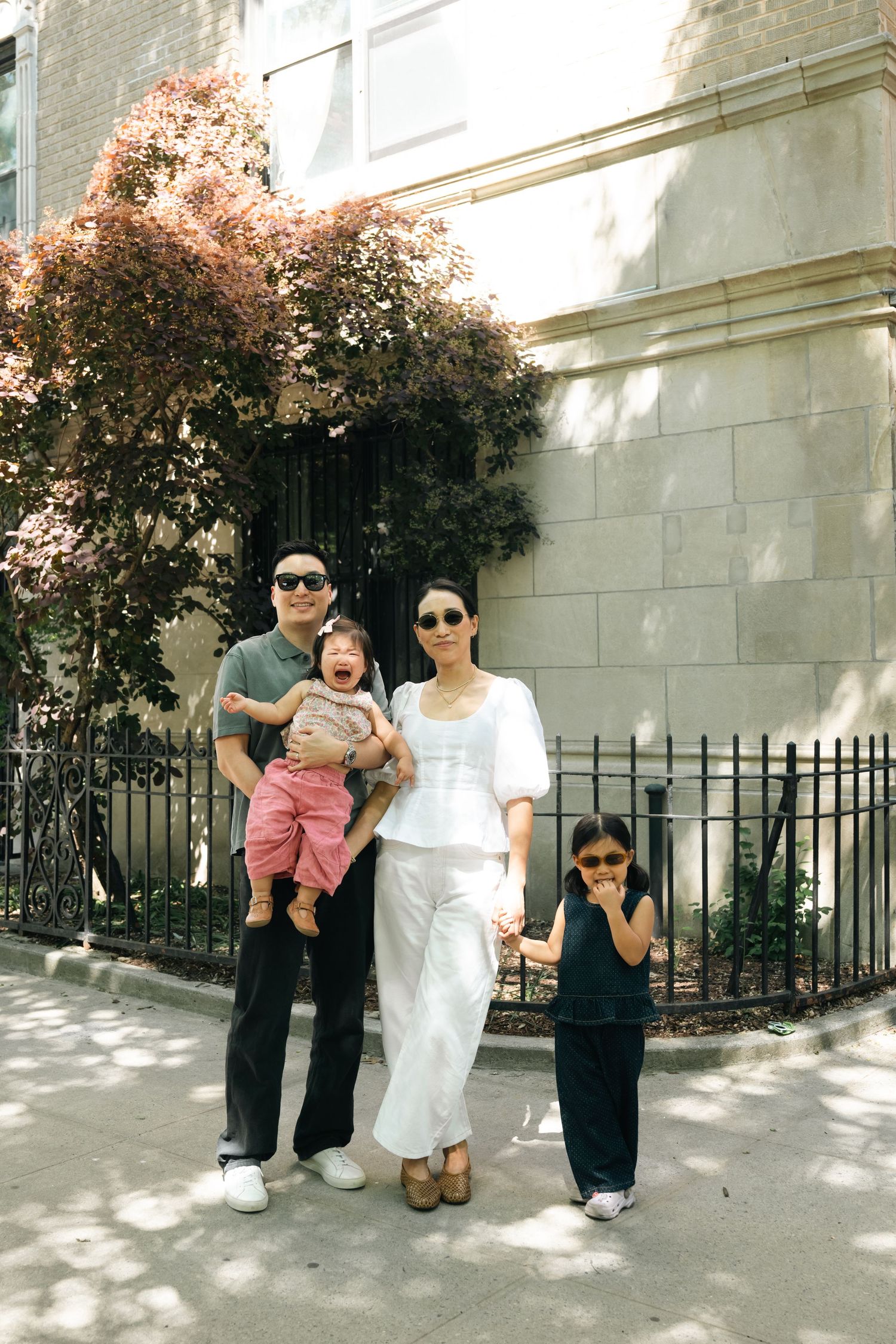 Family of four posing outdoors in front of a stone building with lush trees, wearing sunglasses on a sunny day.