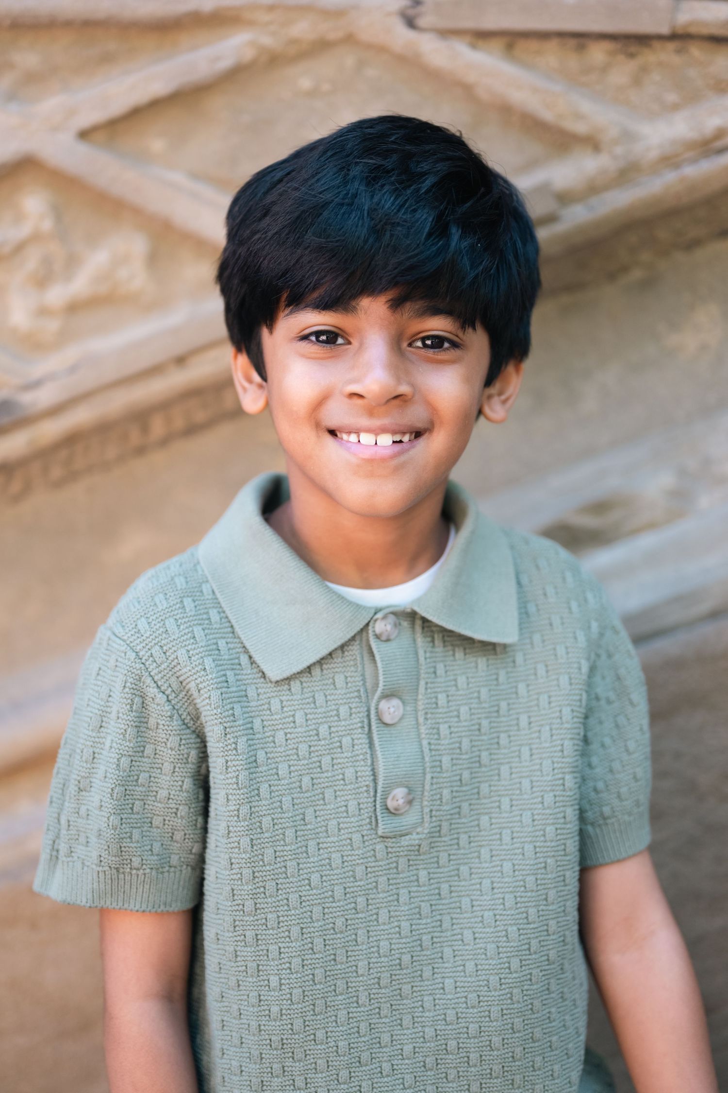 Smiling young boy with black hair wearing a sage green polo shirt standing outdoors near a stone wall.