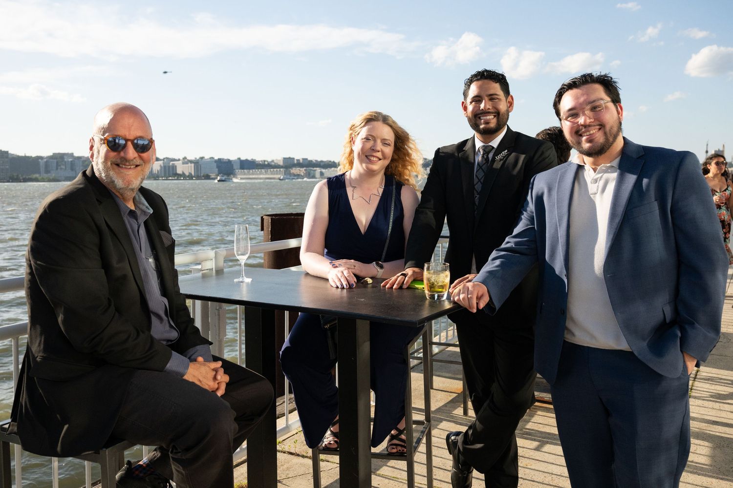 Four elegantly dressed guests pose together at an outdoor waterfront event on a sunny day.