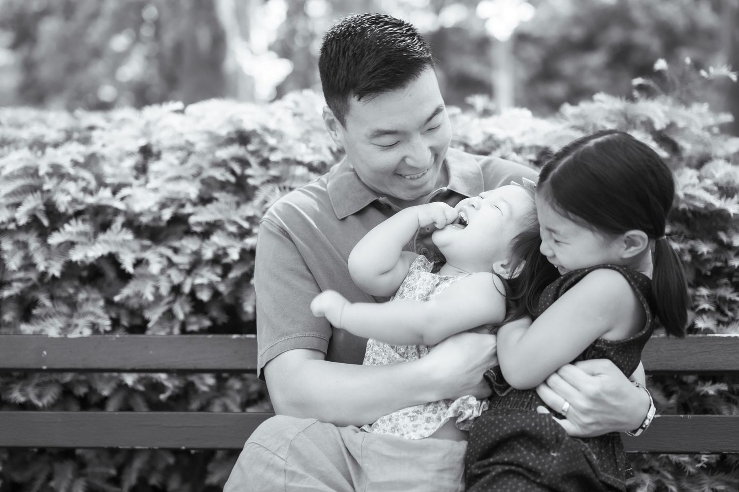 Black and white photo of a smiling Asian father and daughter hugging a laughing baby on a park bench.