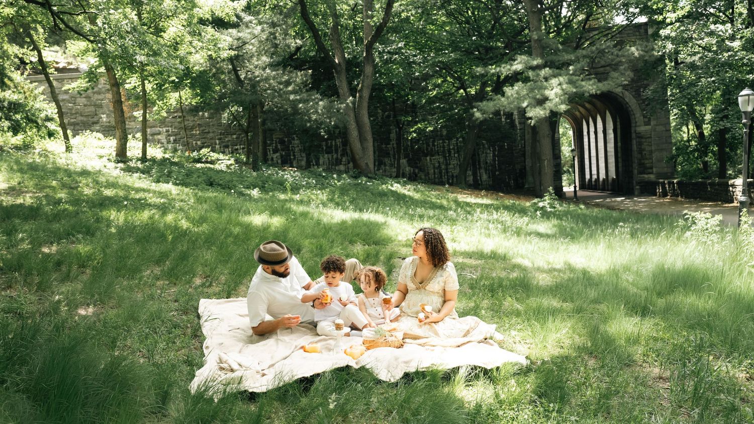 Family enjoying a sunny picnic on a blanket in a lush green park with ancient stone ruins in the background.