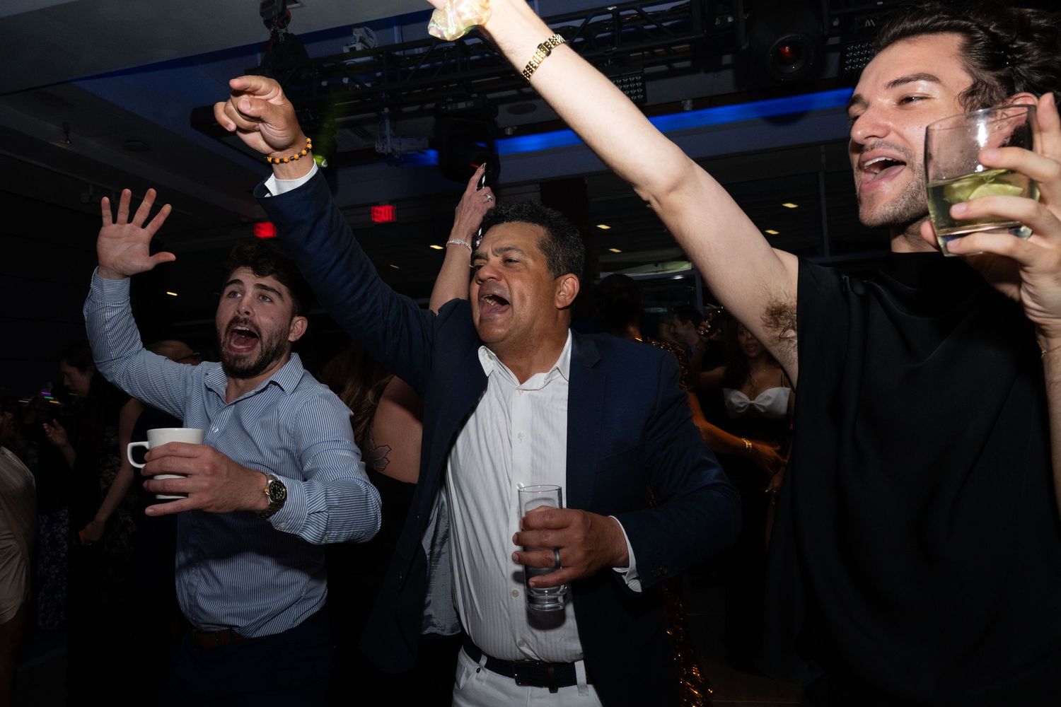 Three men celebrate enthusiastically on a dance floor, raising hands and holding drinks at a party.