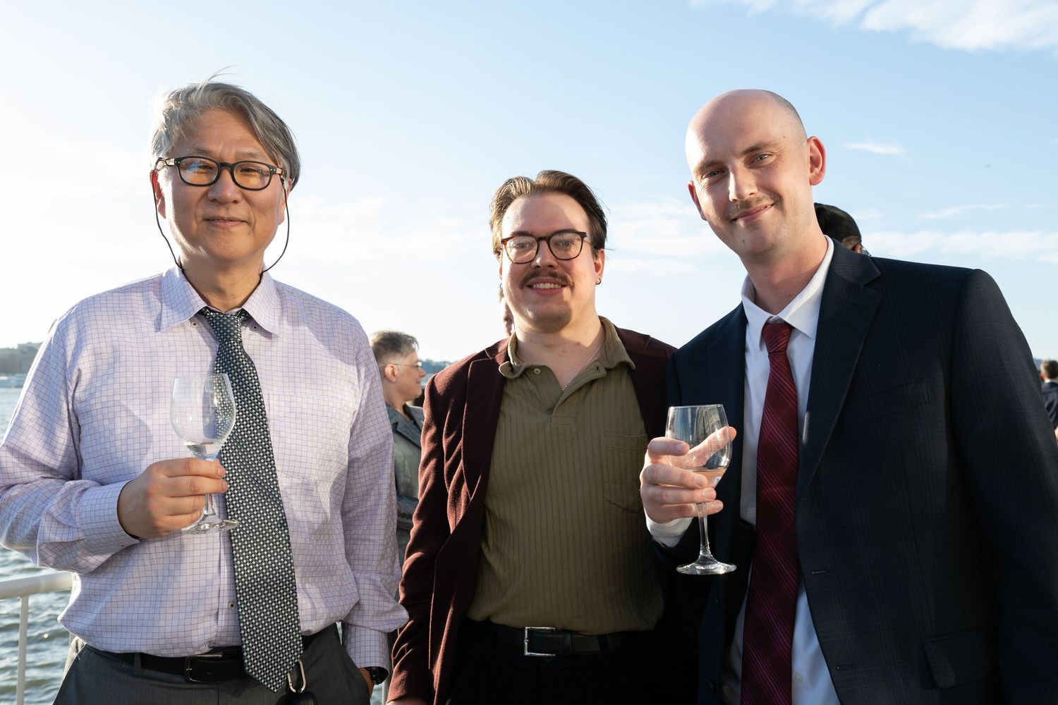 Three men smile and hold wine glasses at an outdoor rooftop event with a blue sky backdrop.