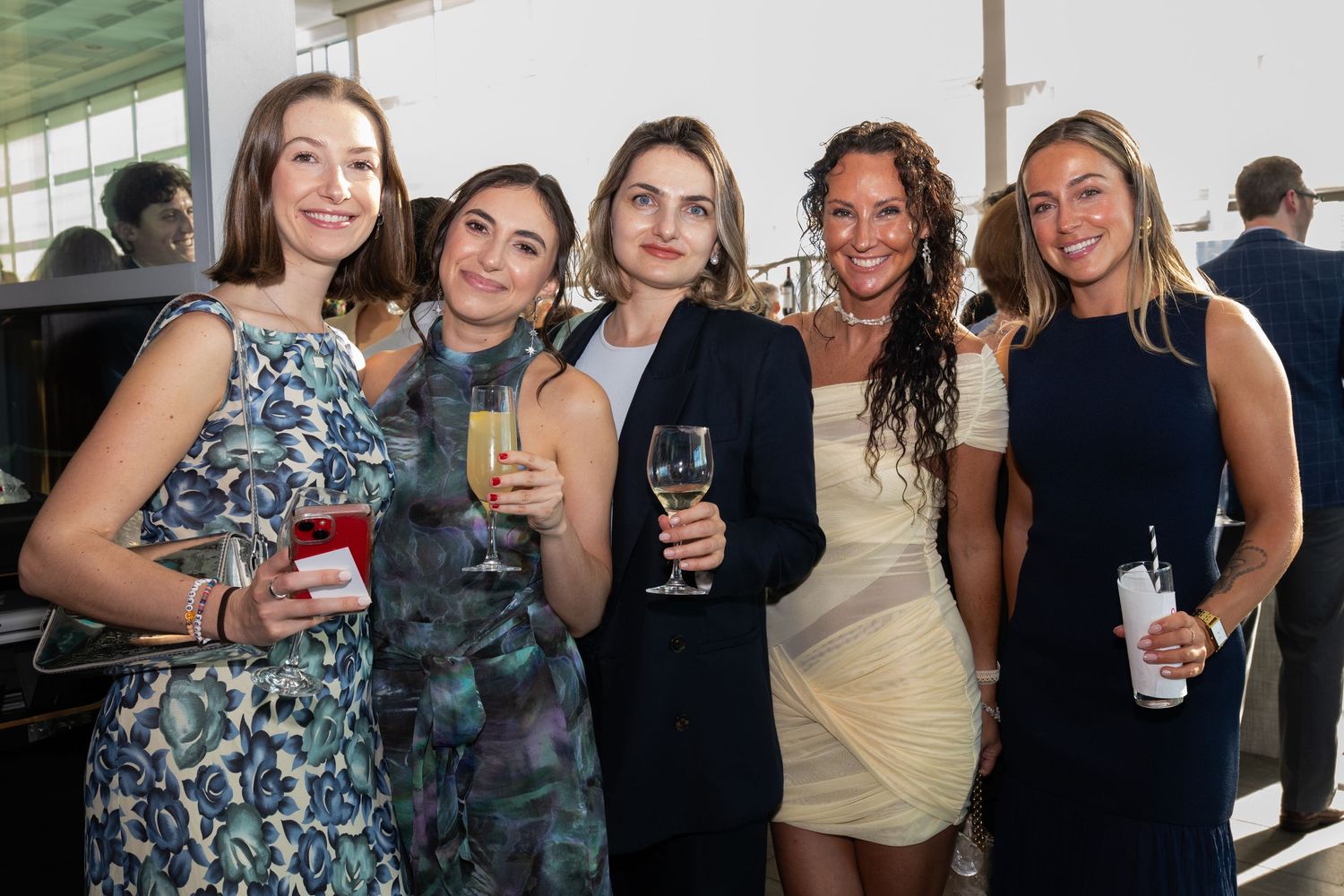 Five women smile and hold drinks at an elegant outdoor social event.