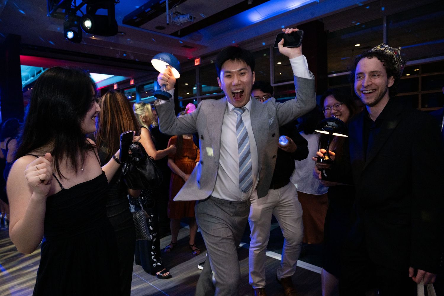 Excited man in gray suit celebrates with raised arms surrounded by dancing guests at a gala party.