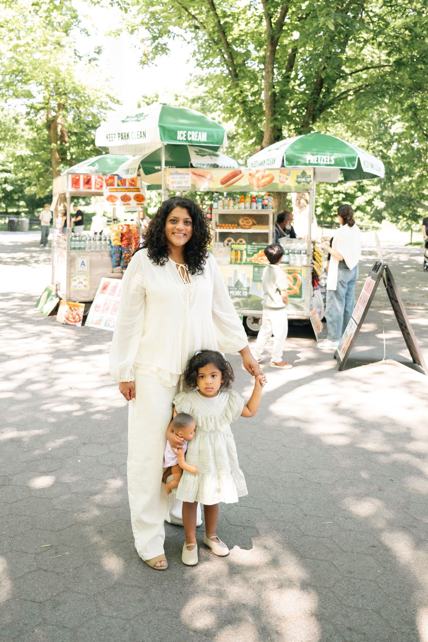 A woman and young girl in white outfits hold hands near a Central Park ice cream cart surrounded by trees.