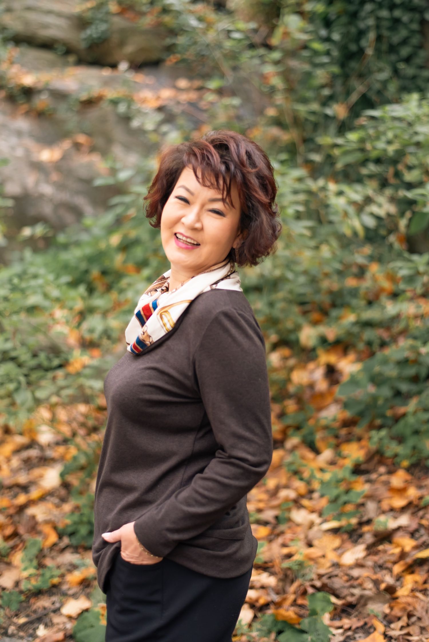 A person in a black sweater poses playfully among autumn leaves in an outdoor setting.
