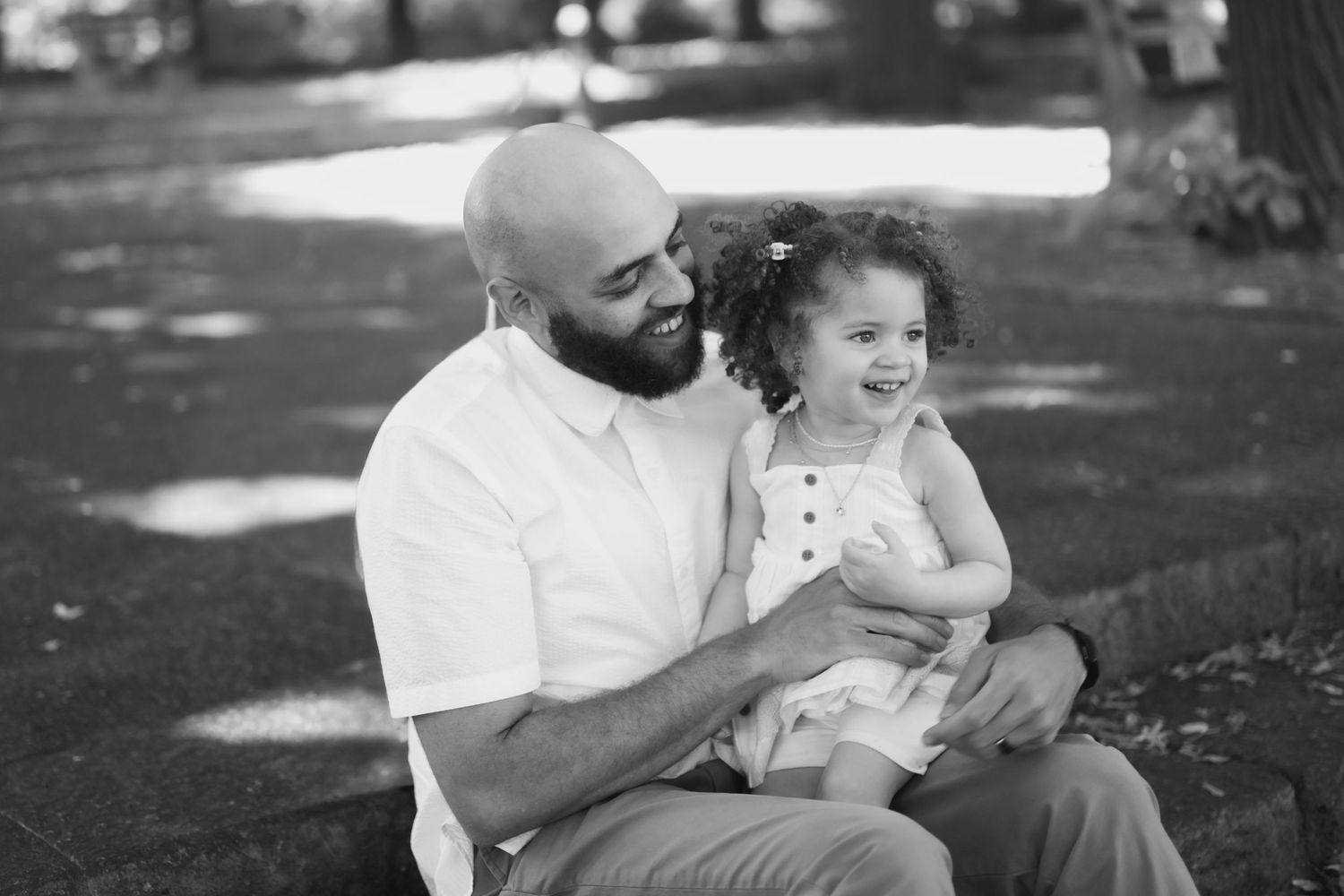 Black and white photo of a bald bearded man smiling at a curly-haired toddler girl sitting on his lap outdoors.