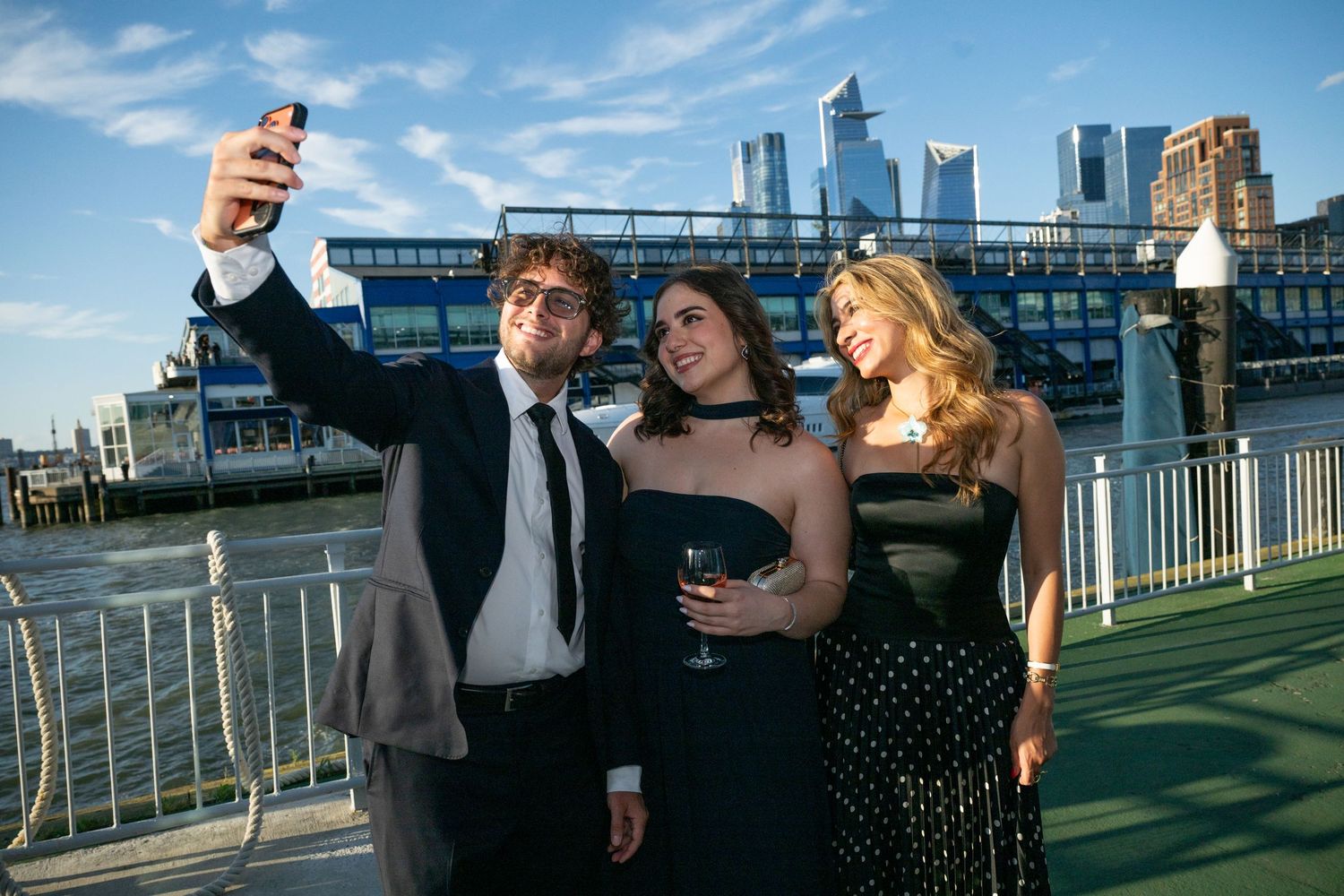 Three elegantly dressed guests take a selfie on a waterfront terrace with a city skyline backdrop at a formal evening event.
