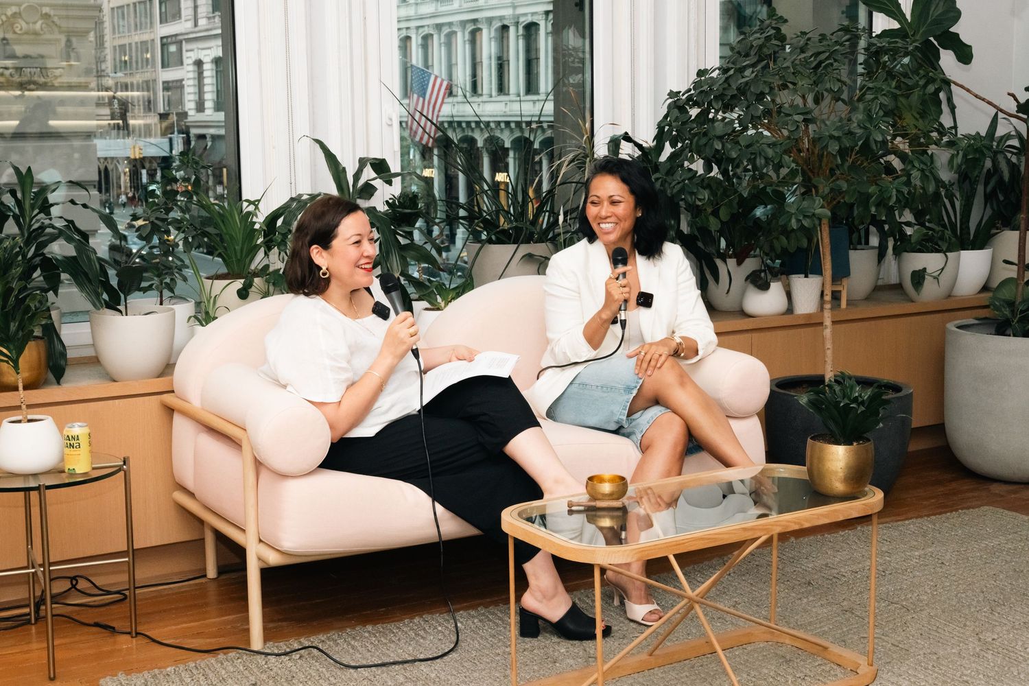 Two women sit on a beige sofa holding microphones, laughing in a plant-filled modern office space.