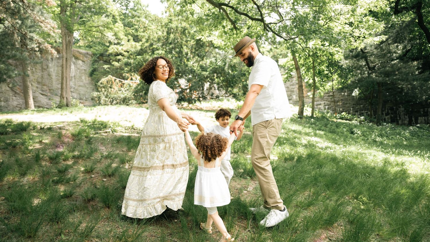A joyful family of four holds hands and spins together on a green lawn surrounded by trees.