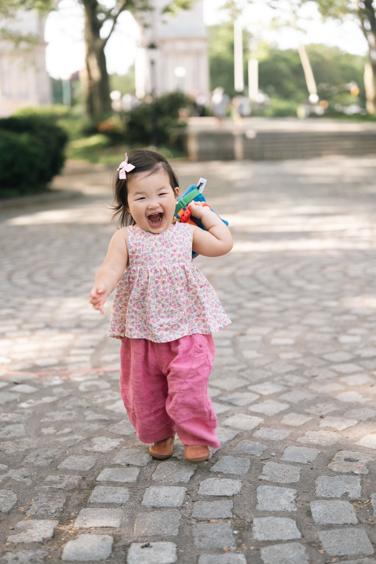 Joyful toddler girl in floral top and pink pants laughing while holding a colorful toy in a sunny park.