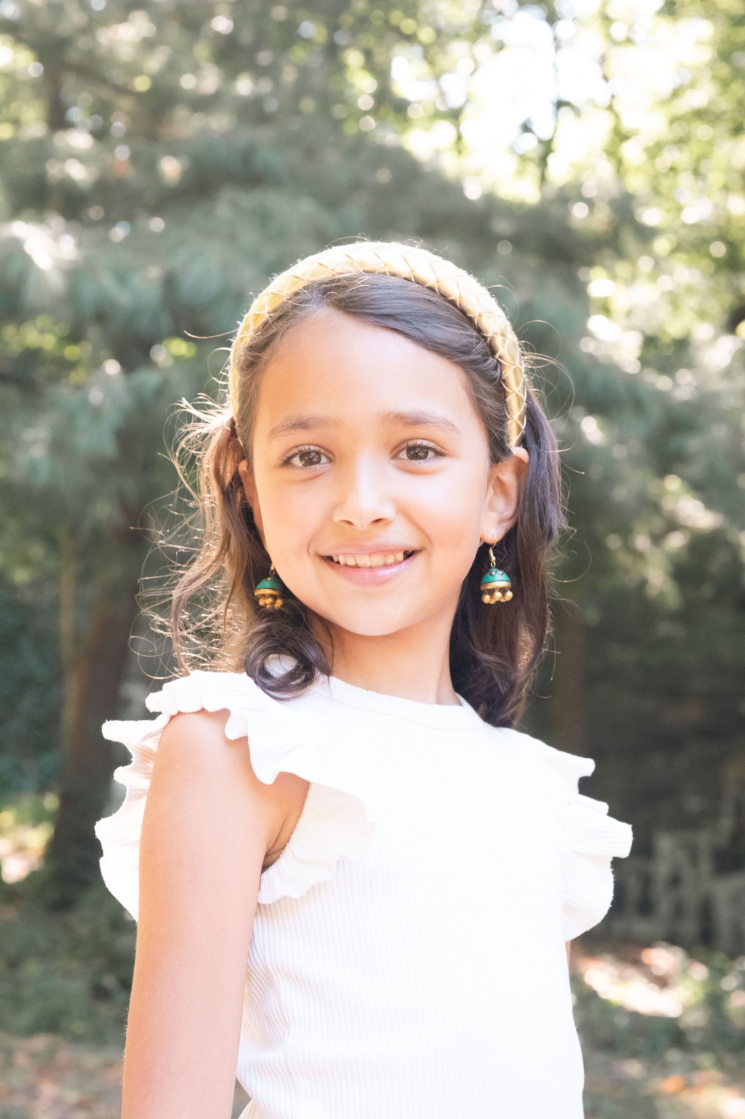 Young girl in white ruffled dress and colorful earrings smiles outdoors with trees in background.