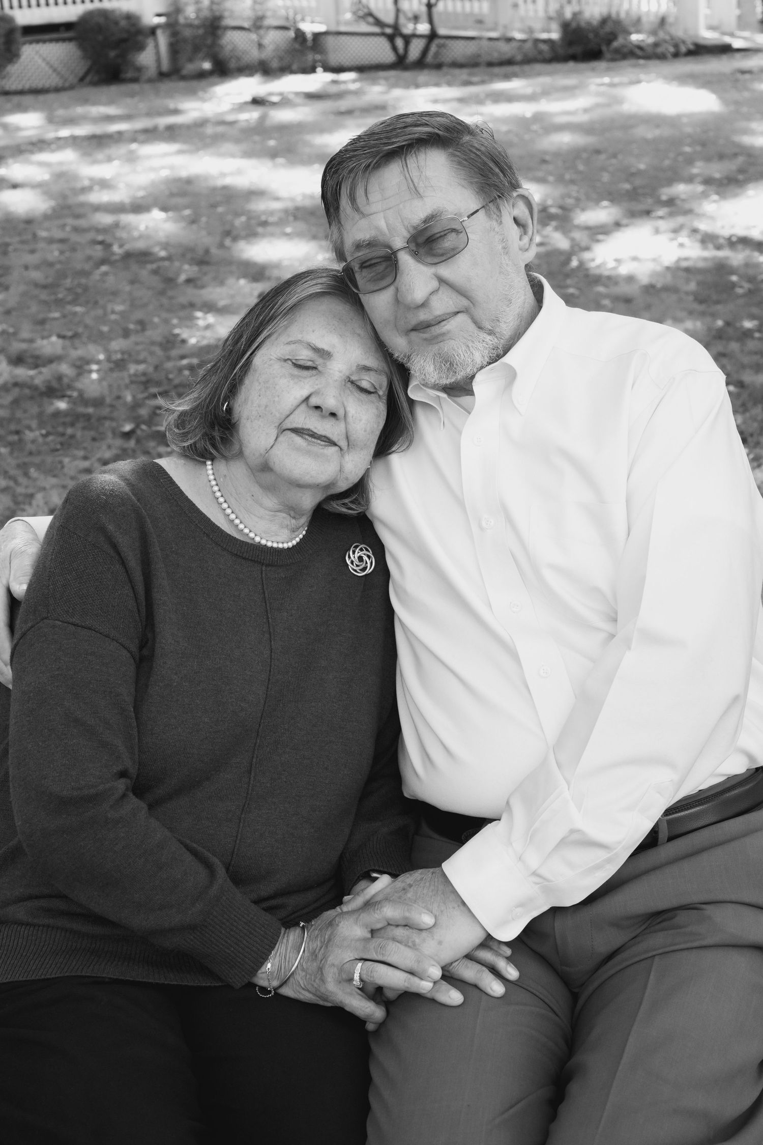 A tender black and white portrait of a couple resting their heads together on a bench.