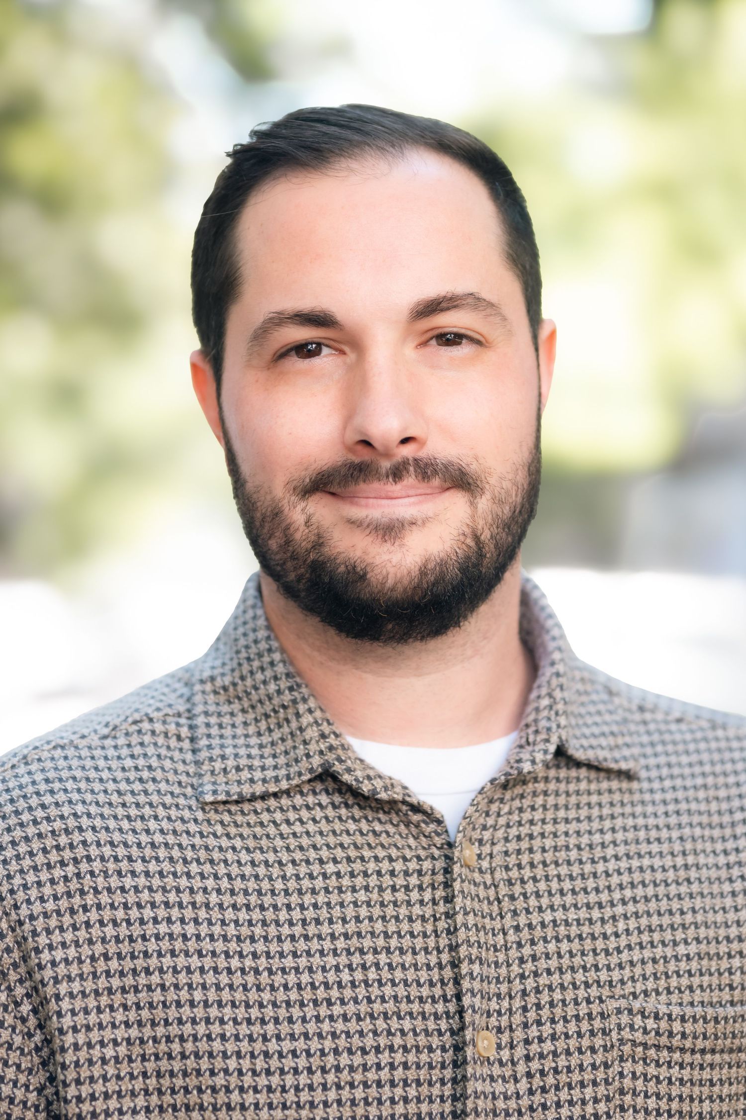 Professional headshot portraits showing warm smile and checkered shirt against blurred background.