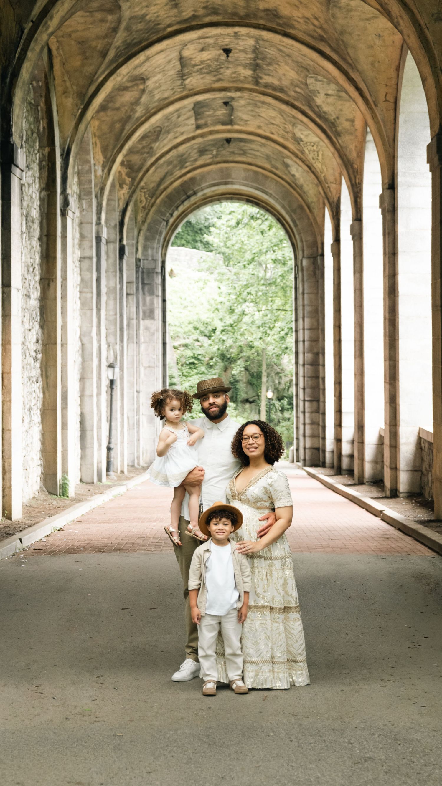 Family of four poses under stone archway with lush greenery, wearing coordinated white and neutral outfits.