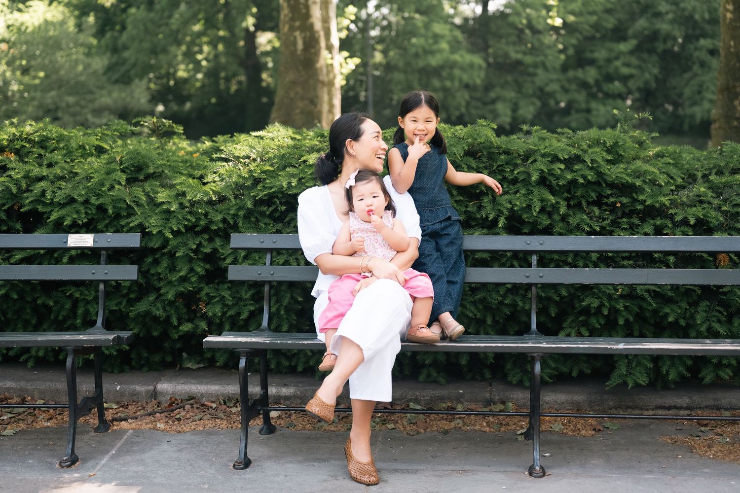 A mother sits on a park bench with two young daughters, one on her lap and one giving her a kiss on the cheek.