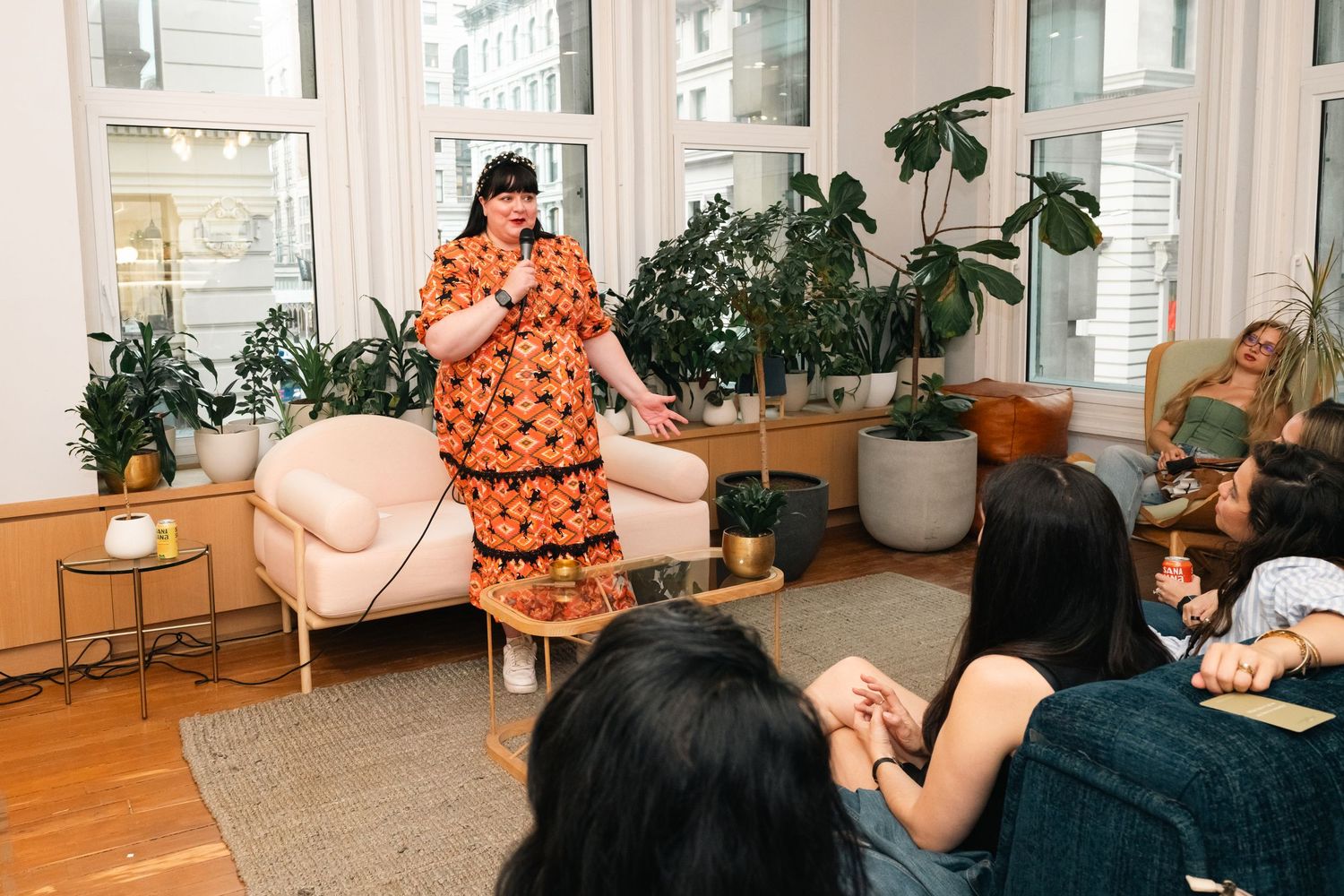 Woman in orange floral dress speaks to seated audience in plant-filled living room during indoor gathering.