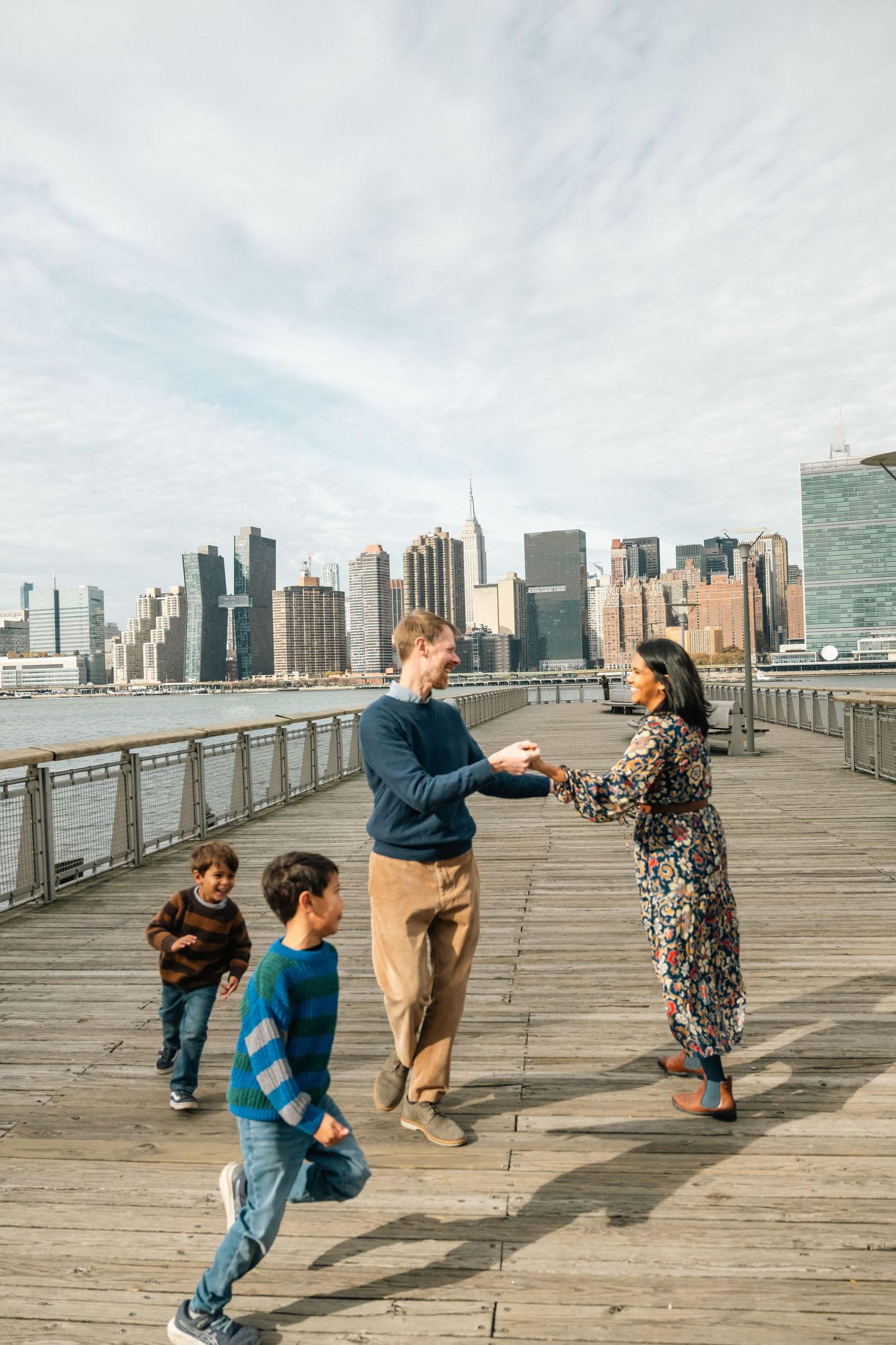Playful family interactions on a wooden boardwalk with Manhattan skyline views.