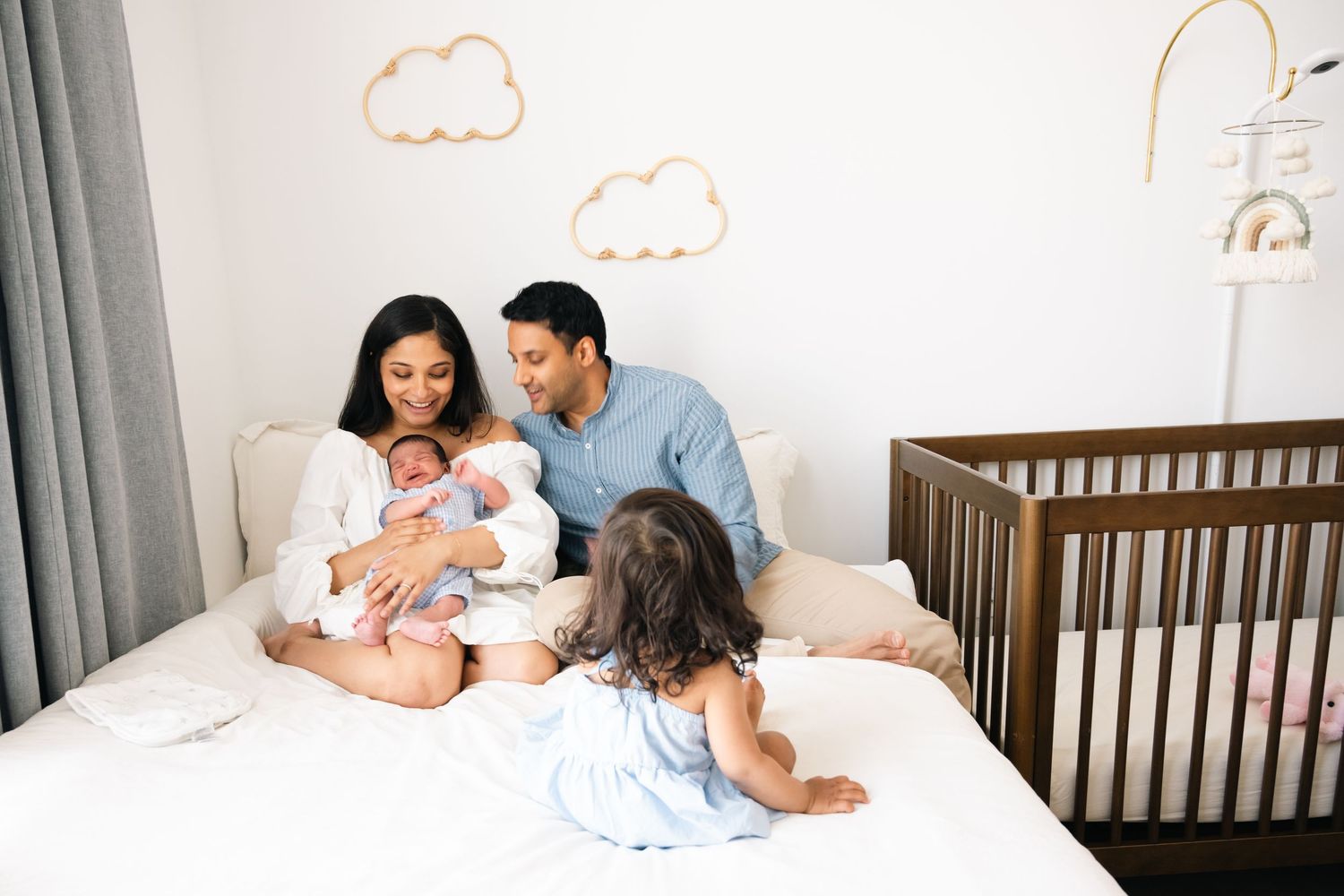 Family relaxing together on white bed with crib and cloud wall decorations in nursery.