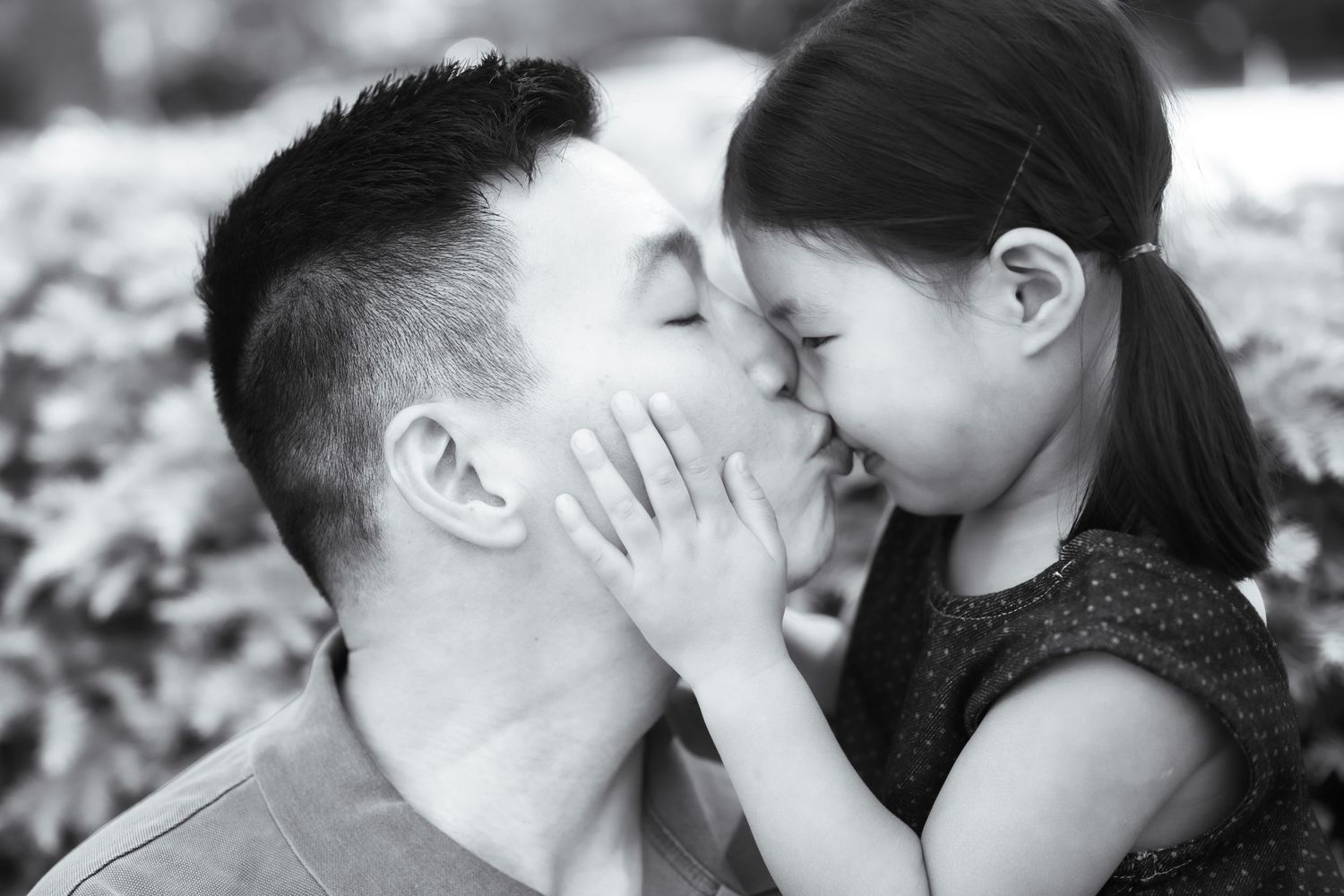 A young girl lovingly kisses a man on the cheek in a tender black and white family portrait outdoors.