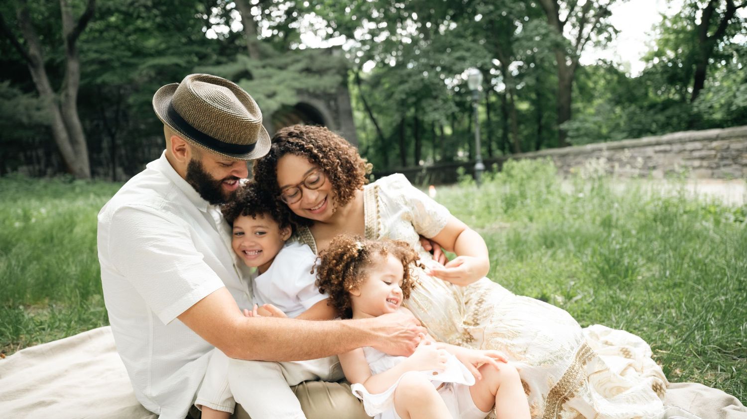 A loving family of four sits together on a blanket in a lush green park, sharing a warm embrace.