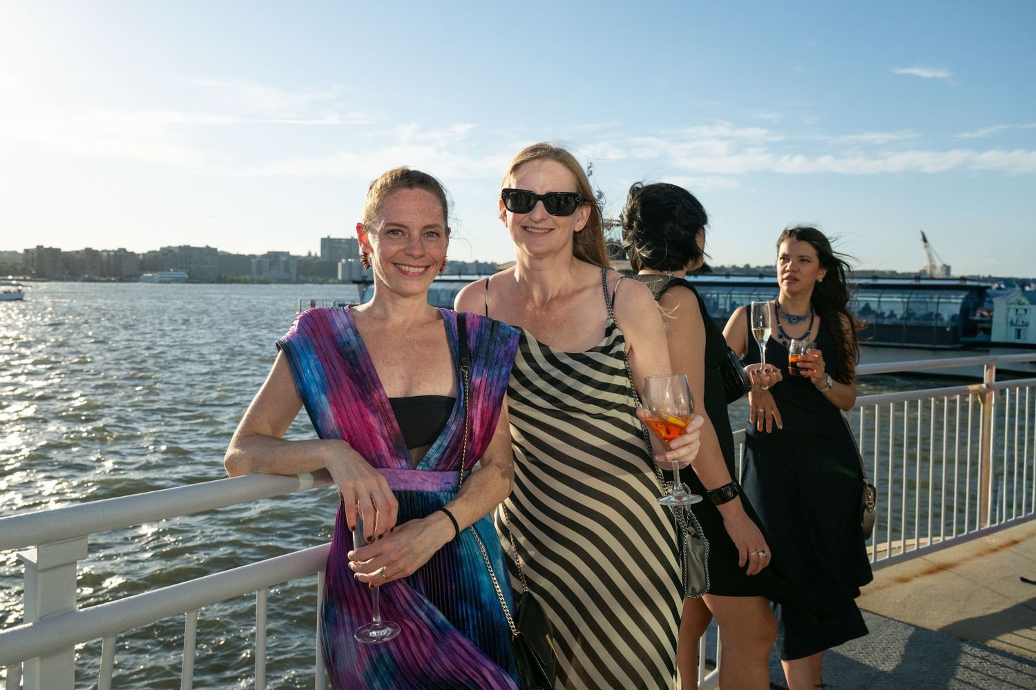 Two women in stylish dresses smile together on a sunny waterfront pier at a social event.