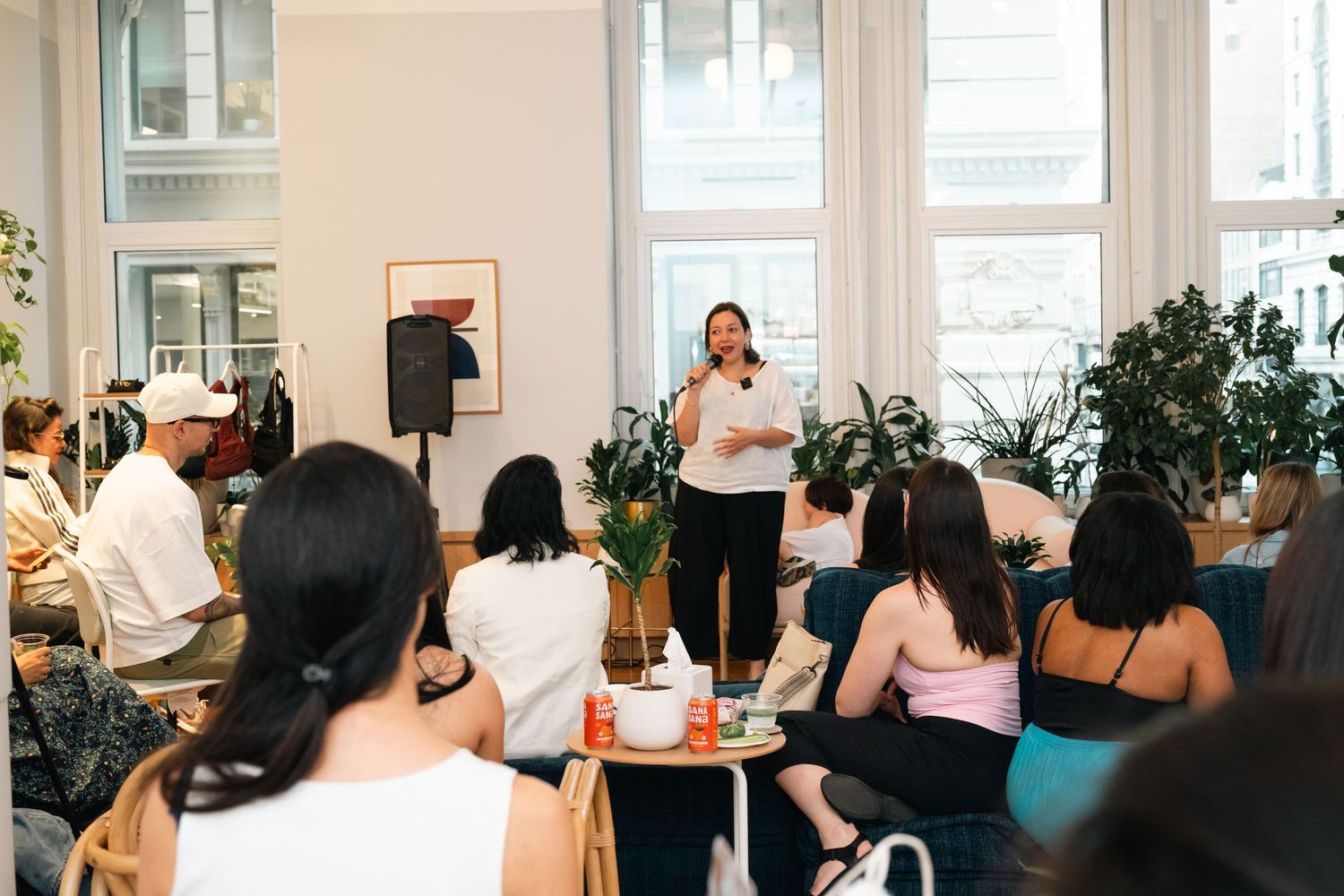 Woman presenting to a diverse seated audience in a bright modern room with plants and large windows.