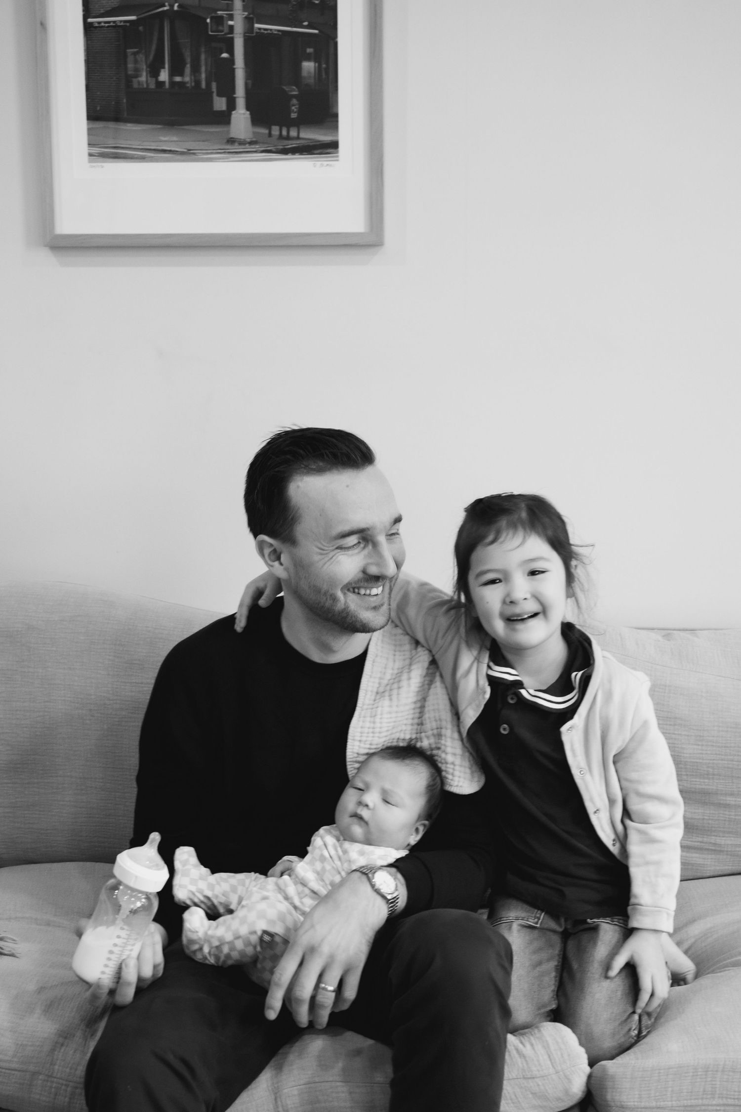 A candid black and white family portrait shows joyful moments shared on a couch with a baby and young child.