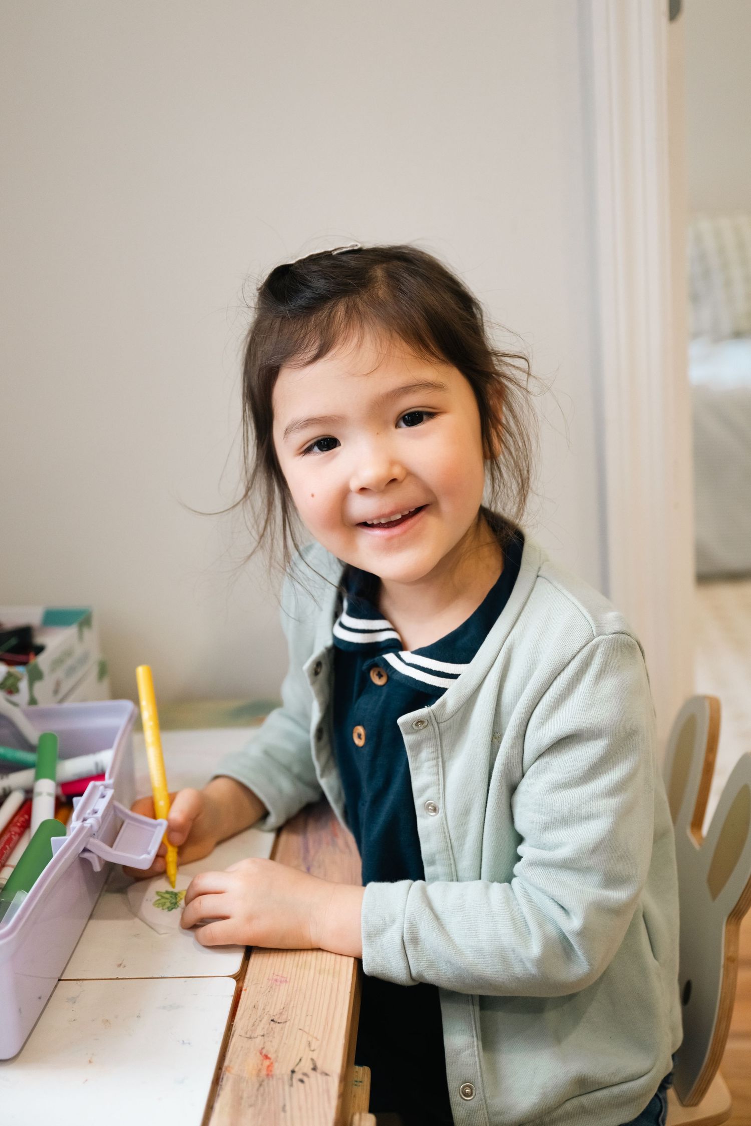 A child sits at a desk smiling while working on homework or schoolwork in a bright indoor setting.