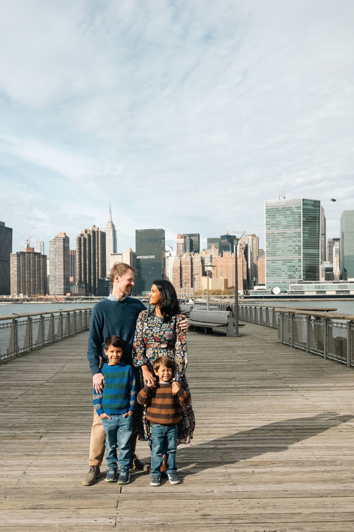 Group walking along wooden pier with New York City skyline views on a cloudy day.
