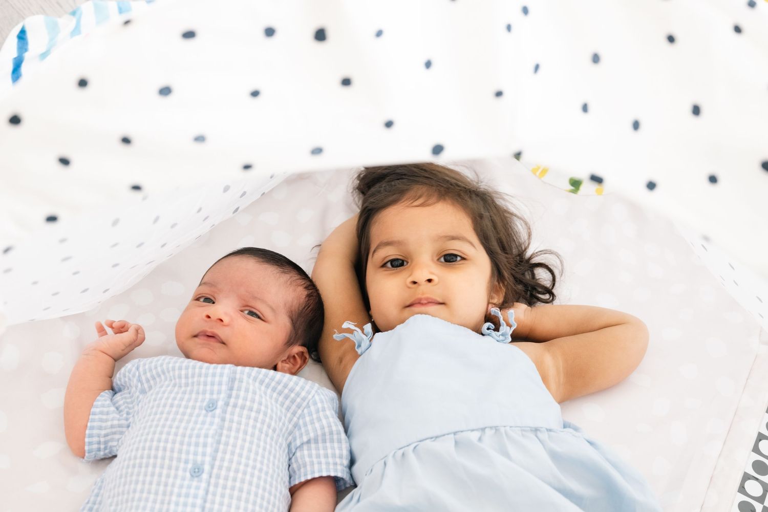Two children lying on polka dot bedding, with newborn beside toddler.