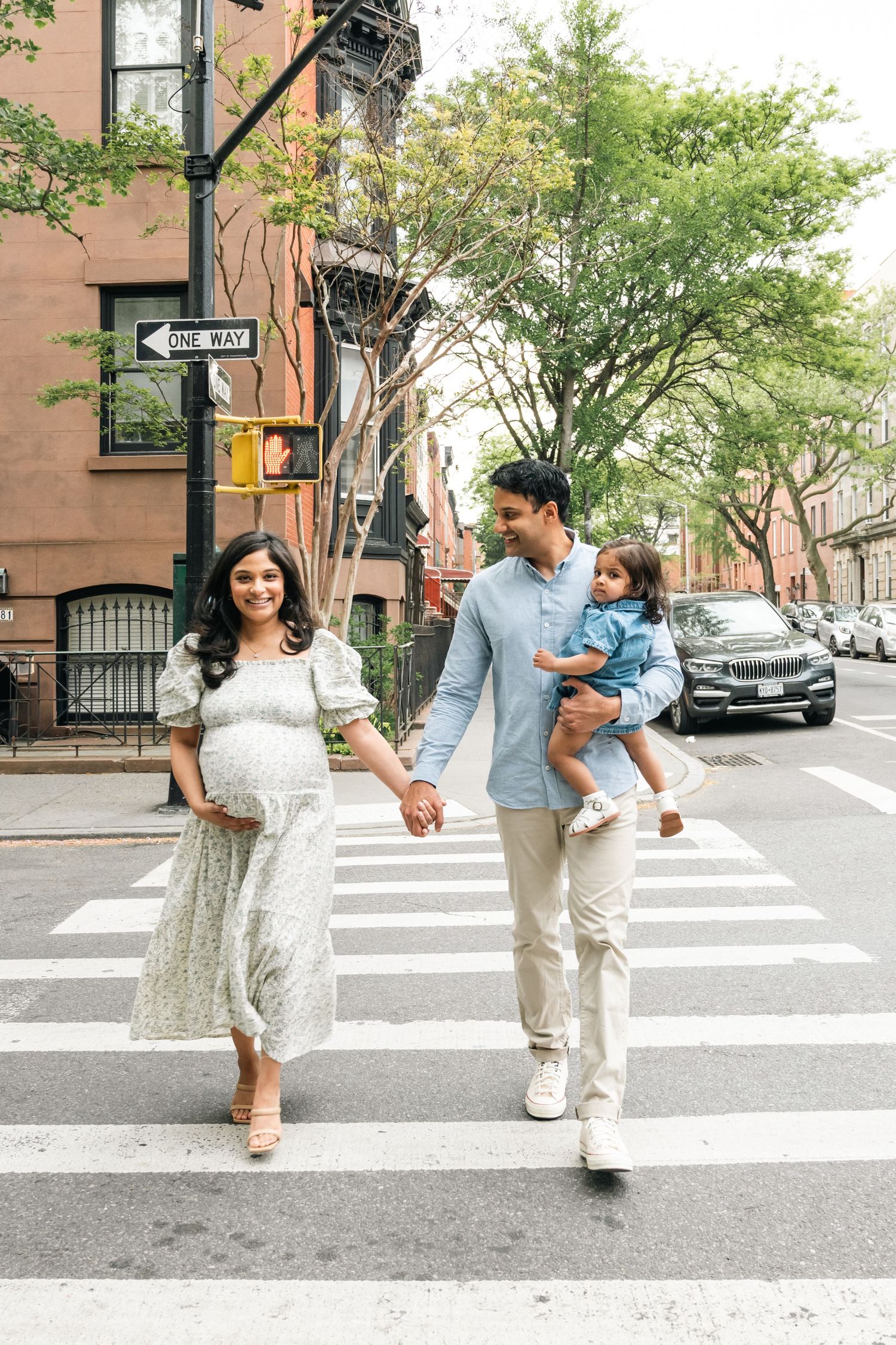 Pregnant woman in floral dress walks with partner carrying toddler across NYC crosswalk on sunny day.