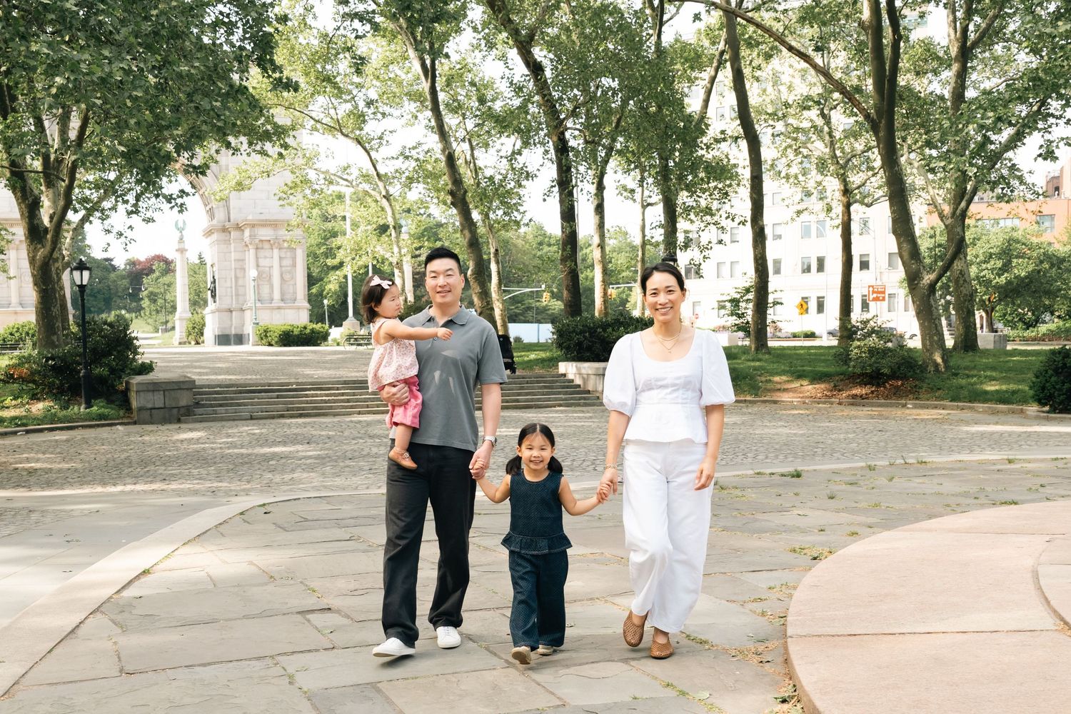 Asian family of four walking in a tree-lined park, parents holding children's hands on a sunny day.