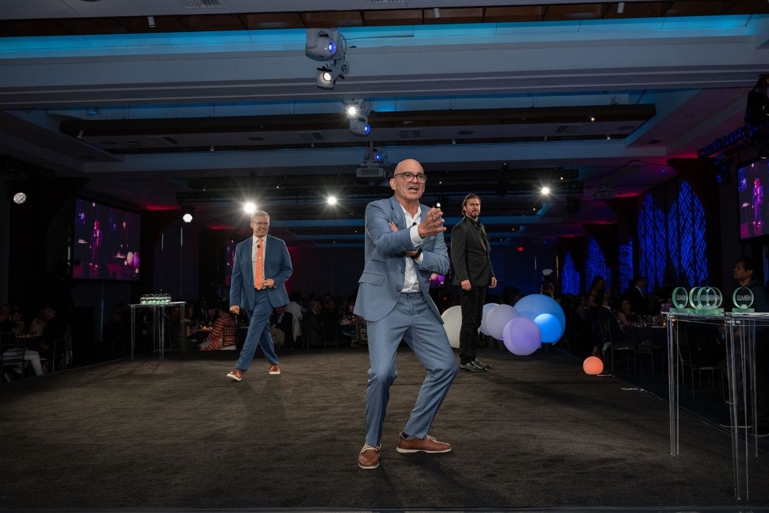 Man in gray suit dances energetically on stage at a corporate gala event with colorful balloons and lighting.