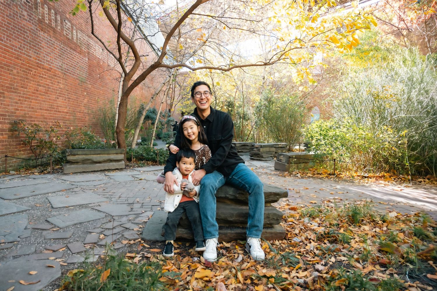 A group sits together on a stone bench surrounded by autumn leaves and brick walls in an outdoor garden setting.