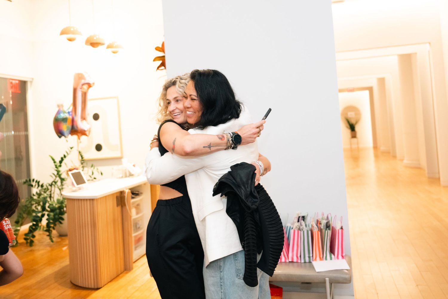 Two women sharing a warm hug inside a bright art gallery, one holding a phone, surrounded by colorful decor.