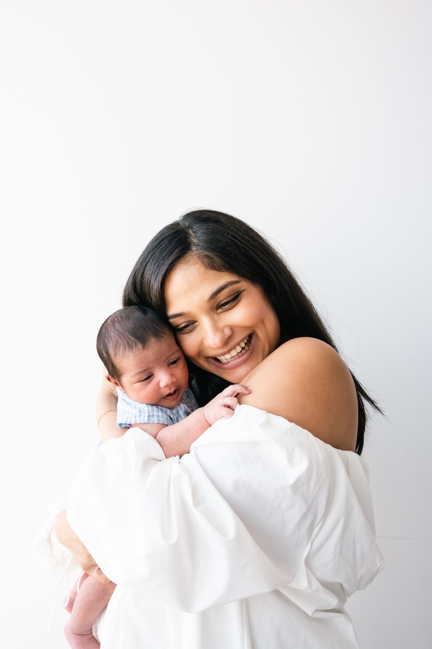 Parent in white off-shoulder top holding sleeping newborn baby against white background.