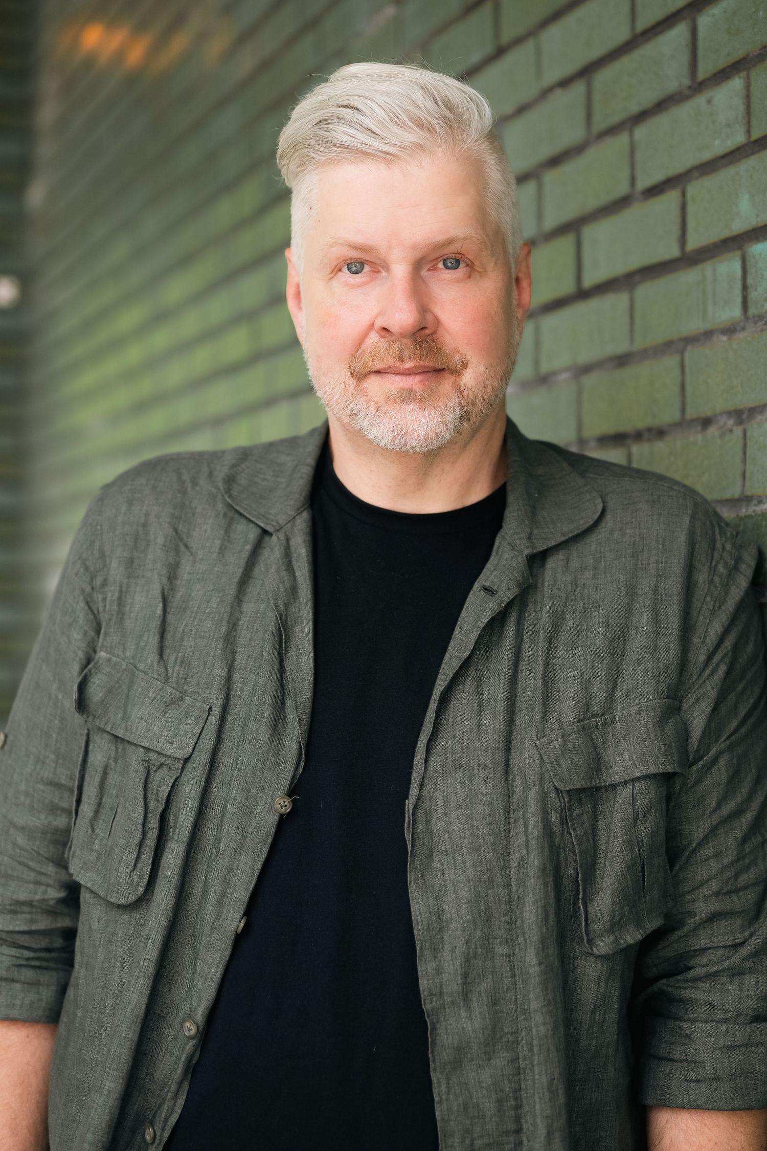 Professional portraits against green brick wall wearing gray jacket and black shirt.