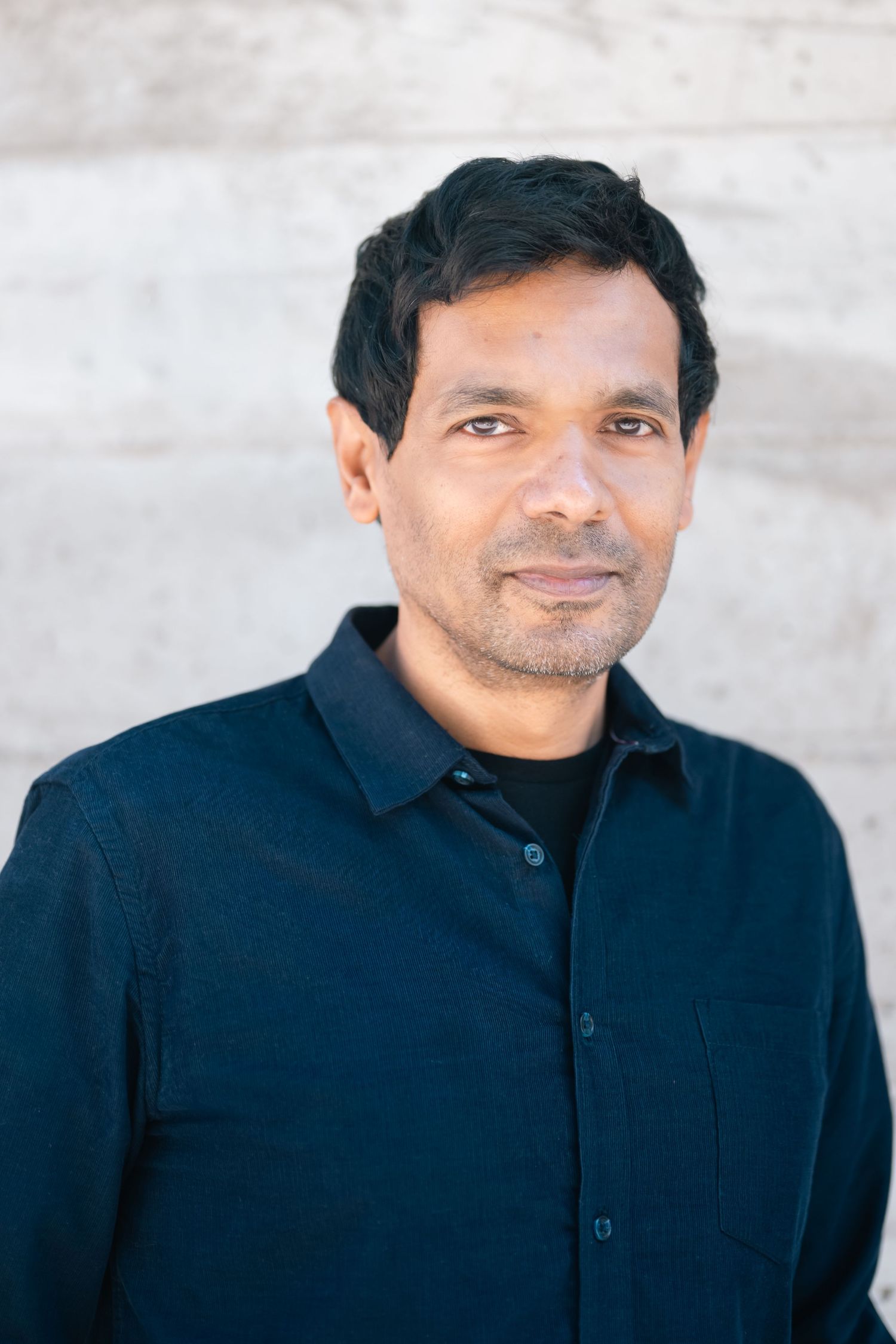 South Asian man with dark hair wearing a navy blue button-up shirt, smiling against a light stone wall.