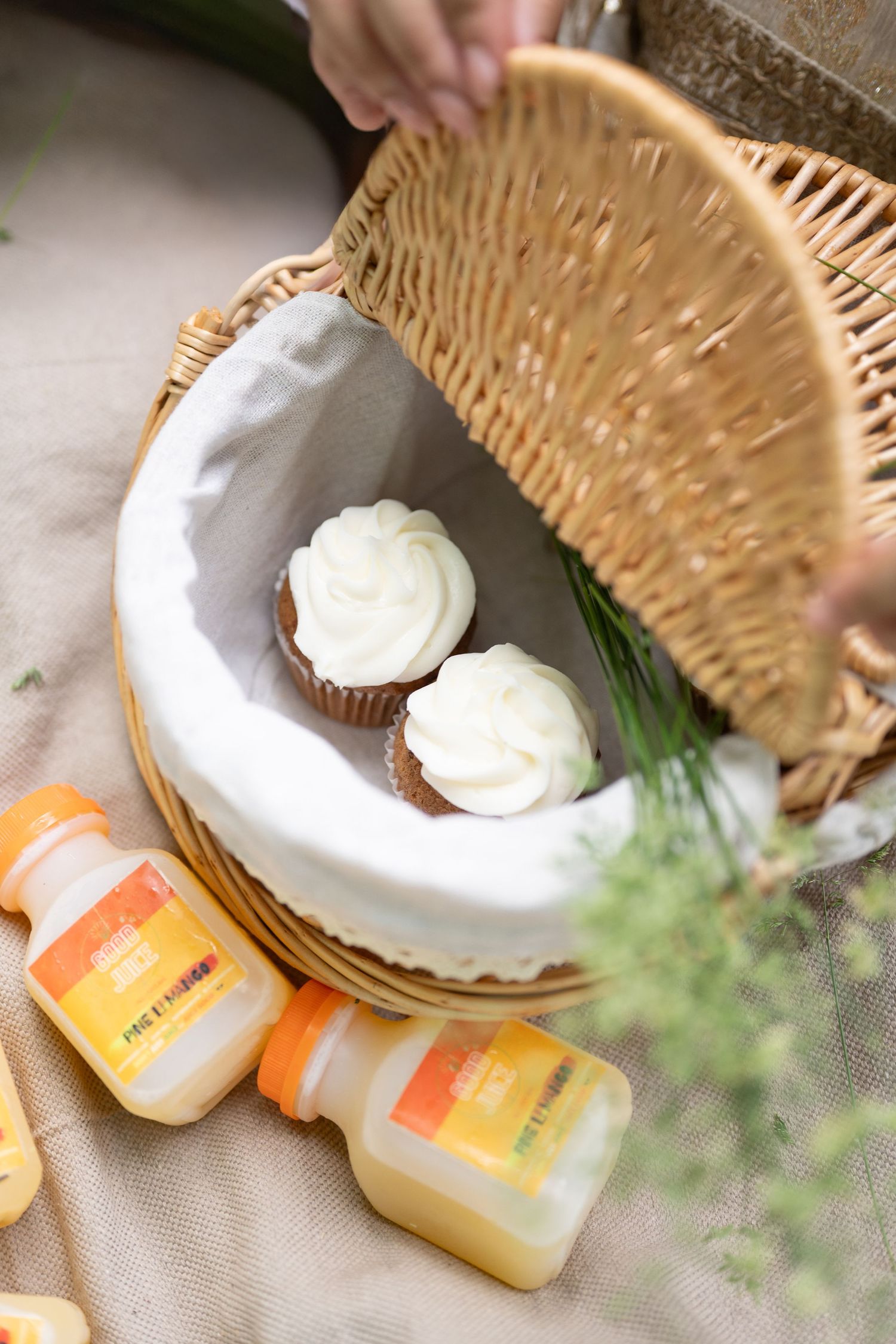 Wicker picnic basket with white-frosted cupcakes inside, surrounded by yellow juice bottles on a linen blanket outdoors.
