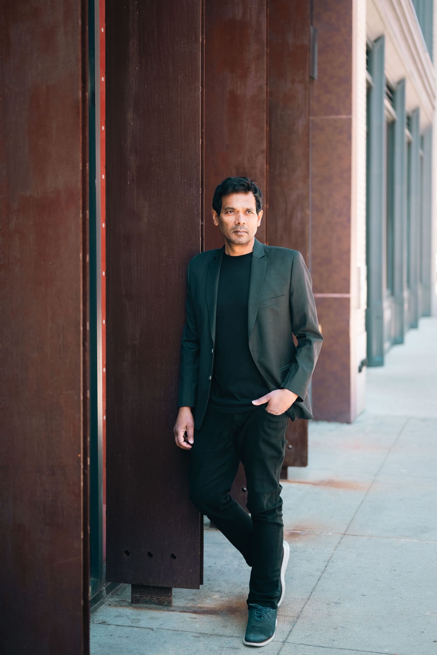 A stylish man in a dark blazer and black outfit leans against a rustic metal wall on an urban sidewalk.