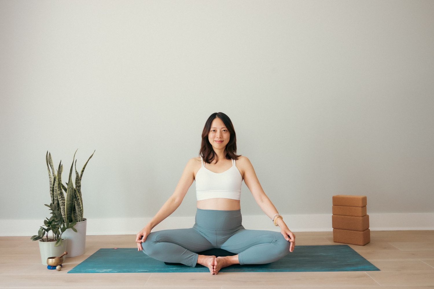 Person in white top and blue leggings practicing yoga on teal mat with potted plant and wooden blocks nearby.