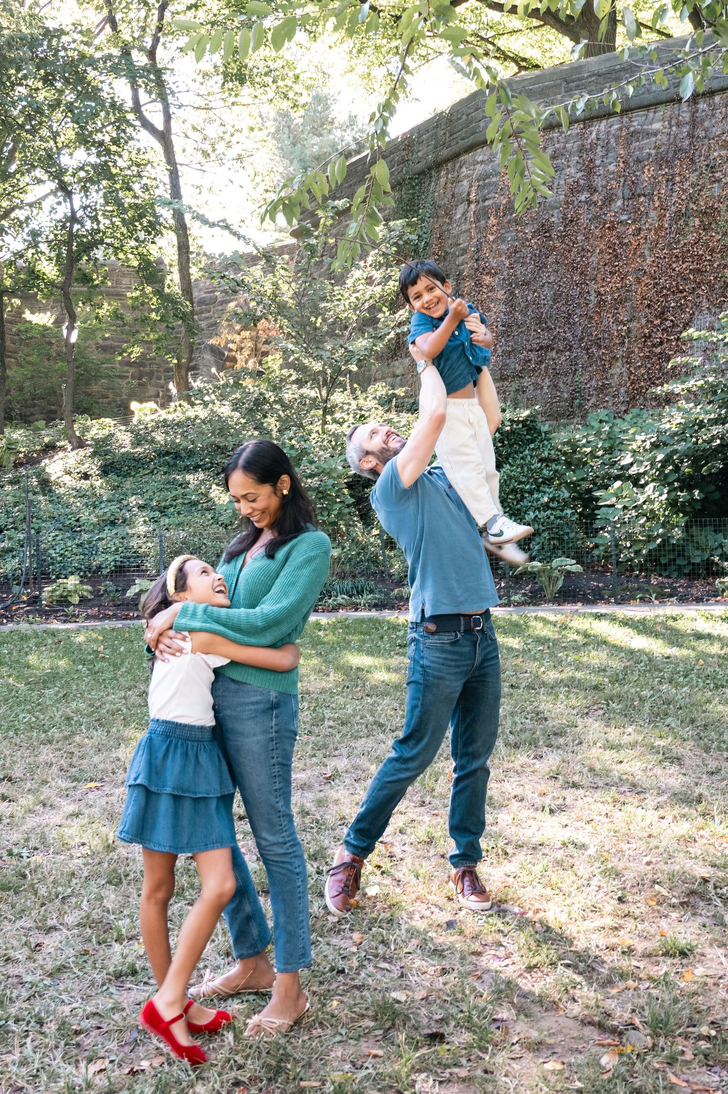A family enjoys quality time together dancing and playing in a garden setting with stone wall and trees.