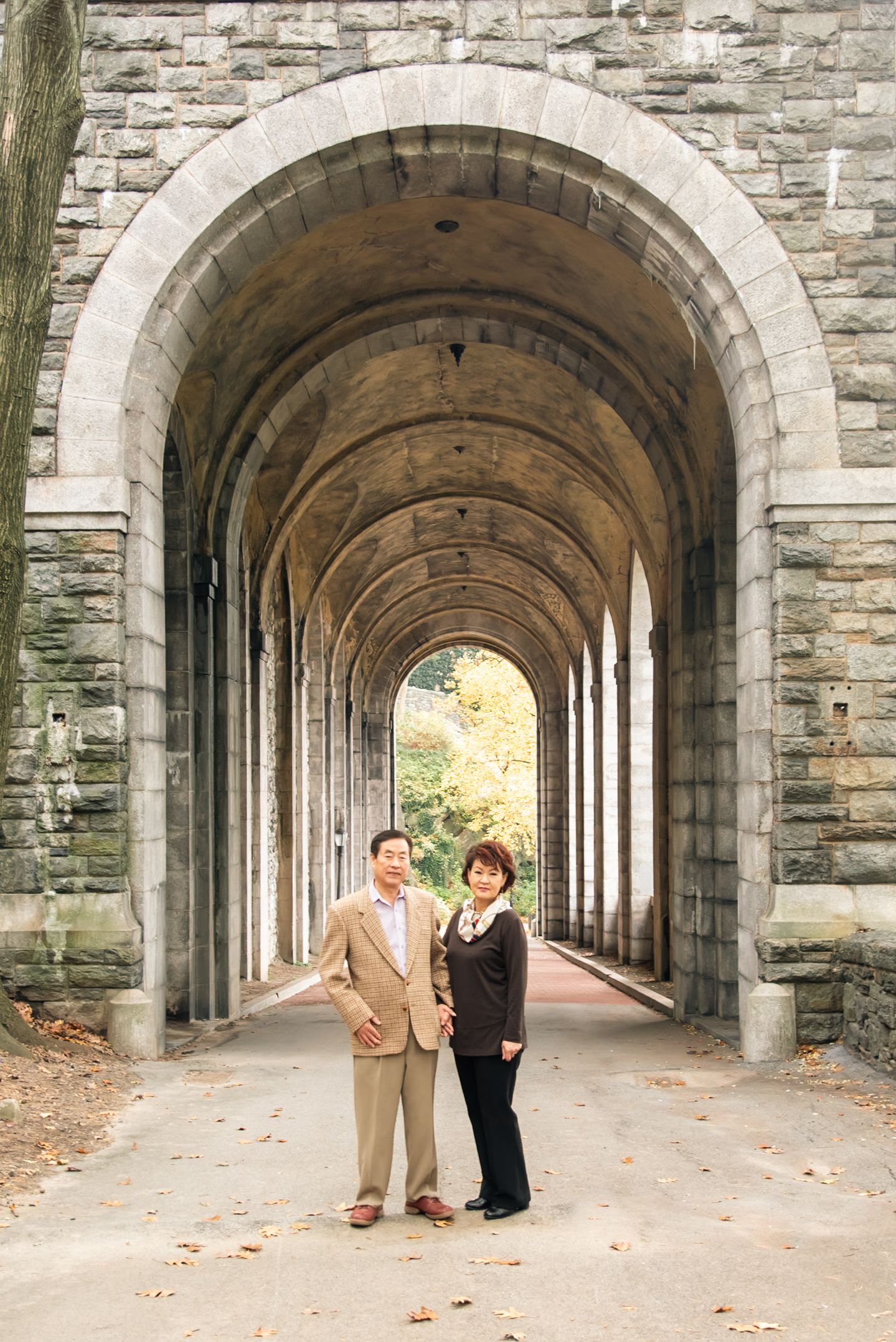 Two people pose together under a beautiful stone archway corridor with natural lighting.