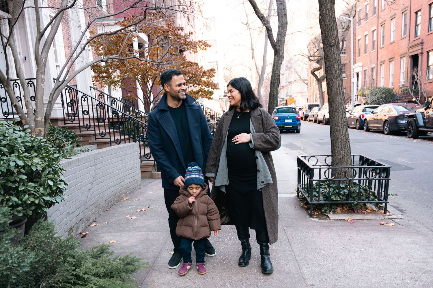 A happy family of three walks on a tree-lined urban sidewalk, with a pregnant mother, father, and young child.