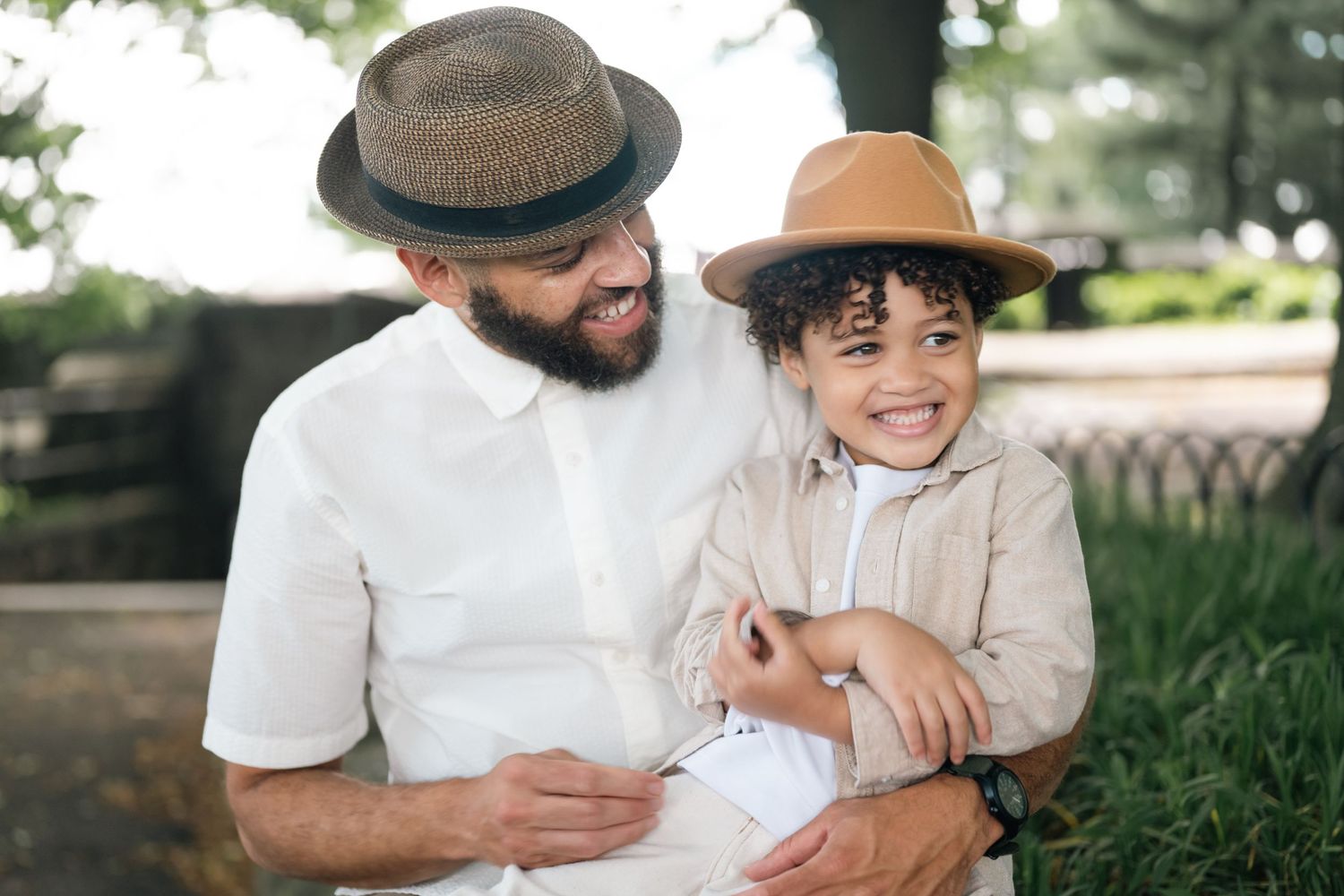 A bearded man and young boy wearing matching fedora hats smile together outdoors in a park.