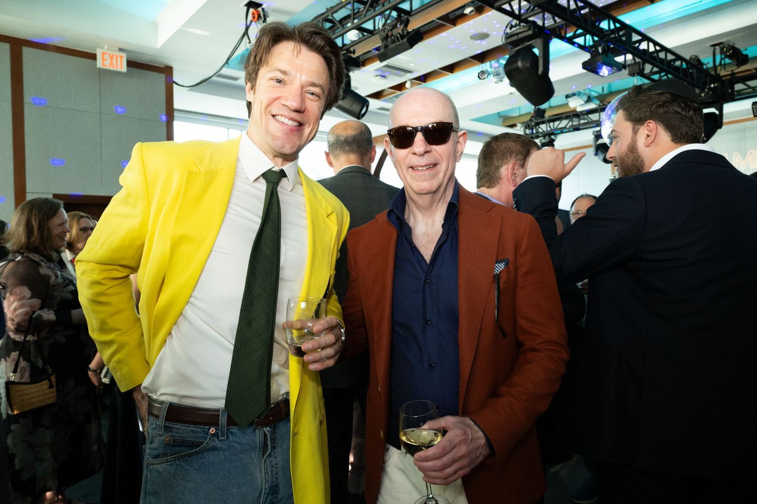 Two men smile together at a gala, one wearing a bold yellow blazer and the other a rust-colored jacket.
