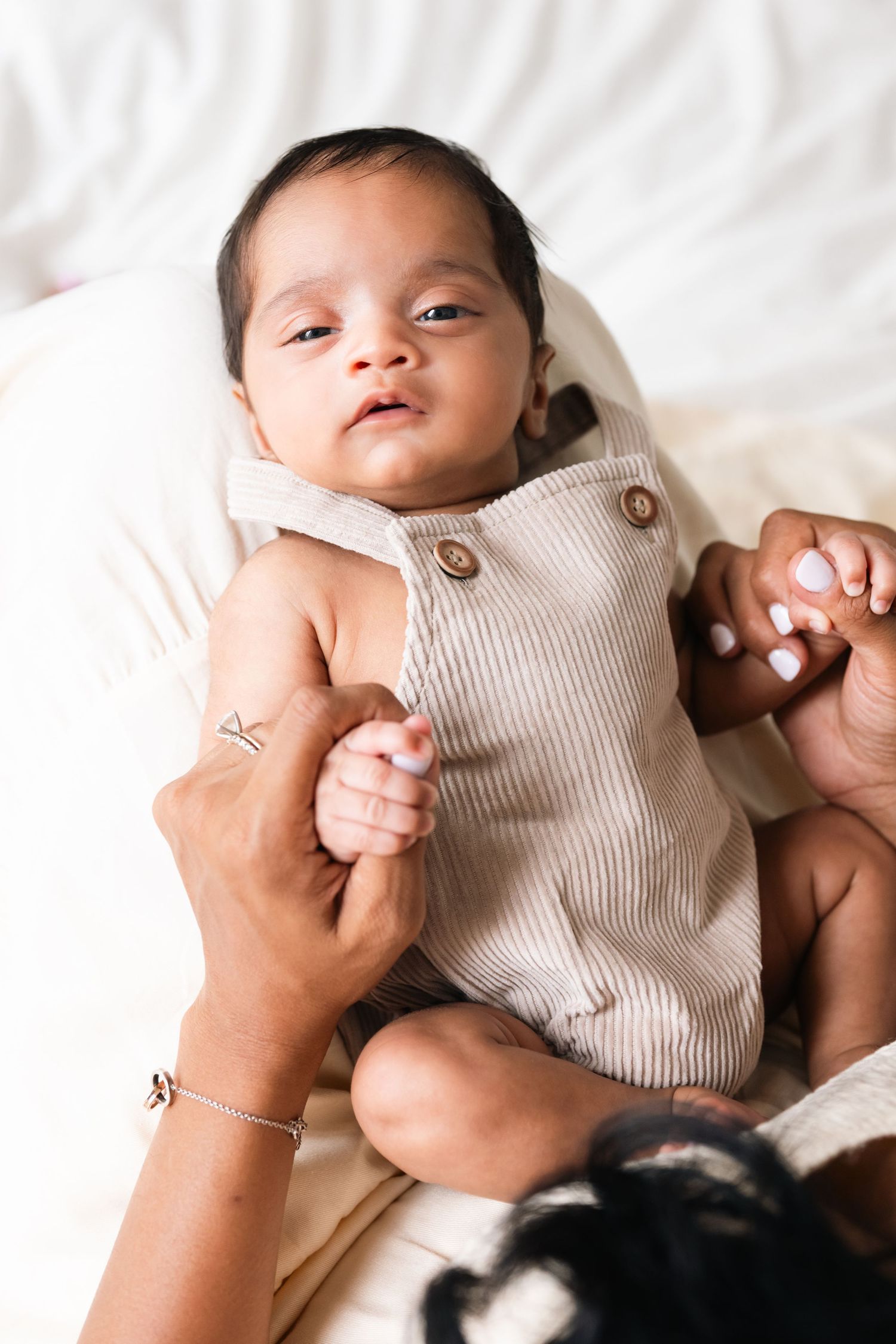 Baby wearing beige romper lying on white bedding while being gently held.