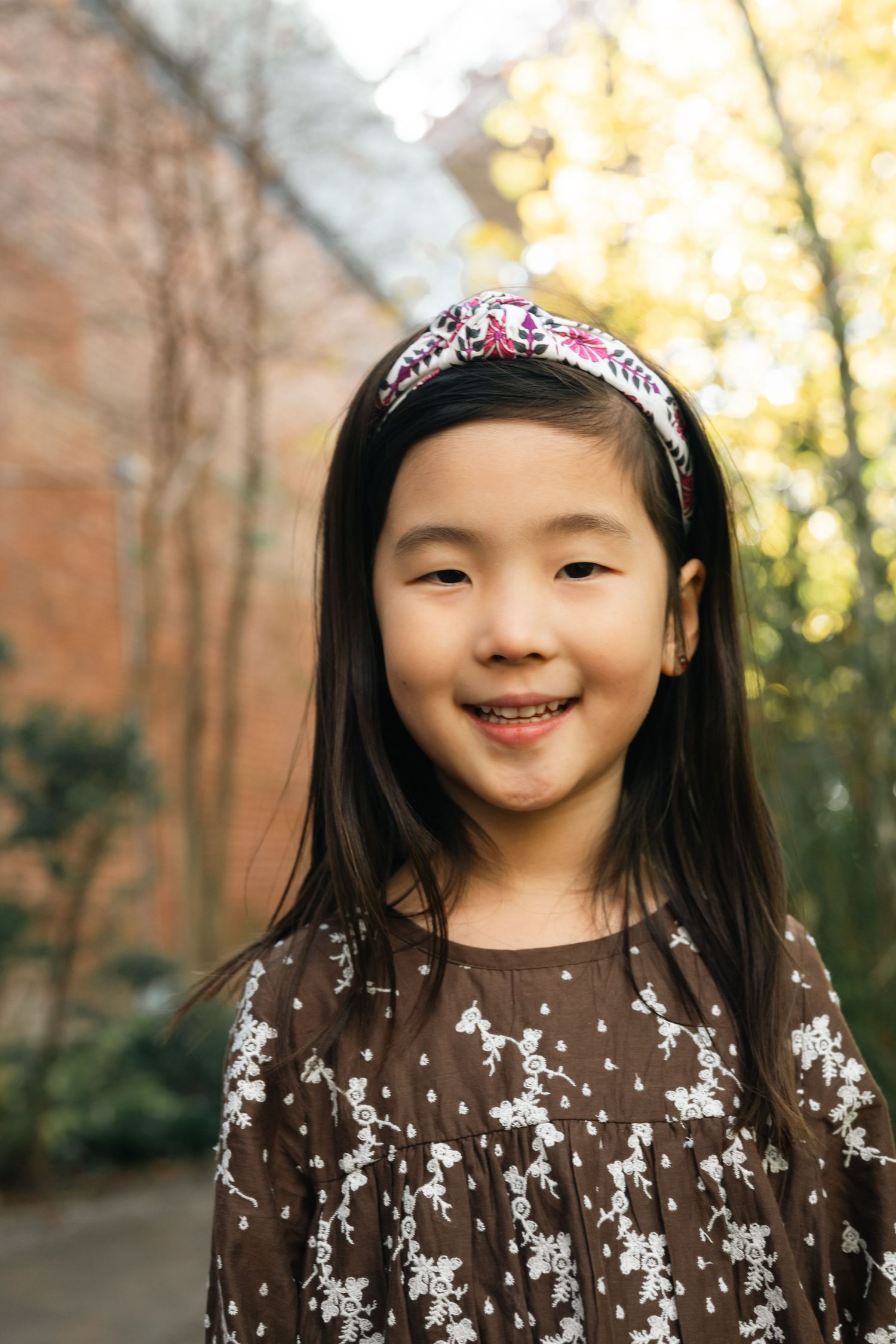 A child wearing a floral headband and brown sweater smiles joyfully outdoors with autumn trees in the background.